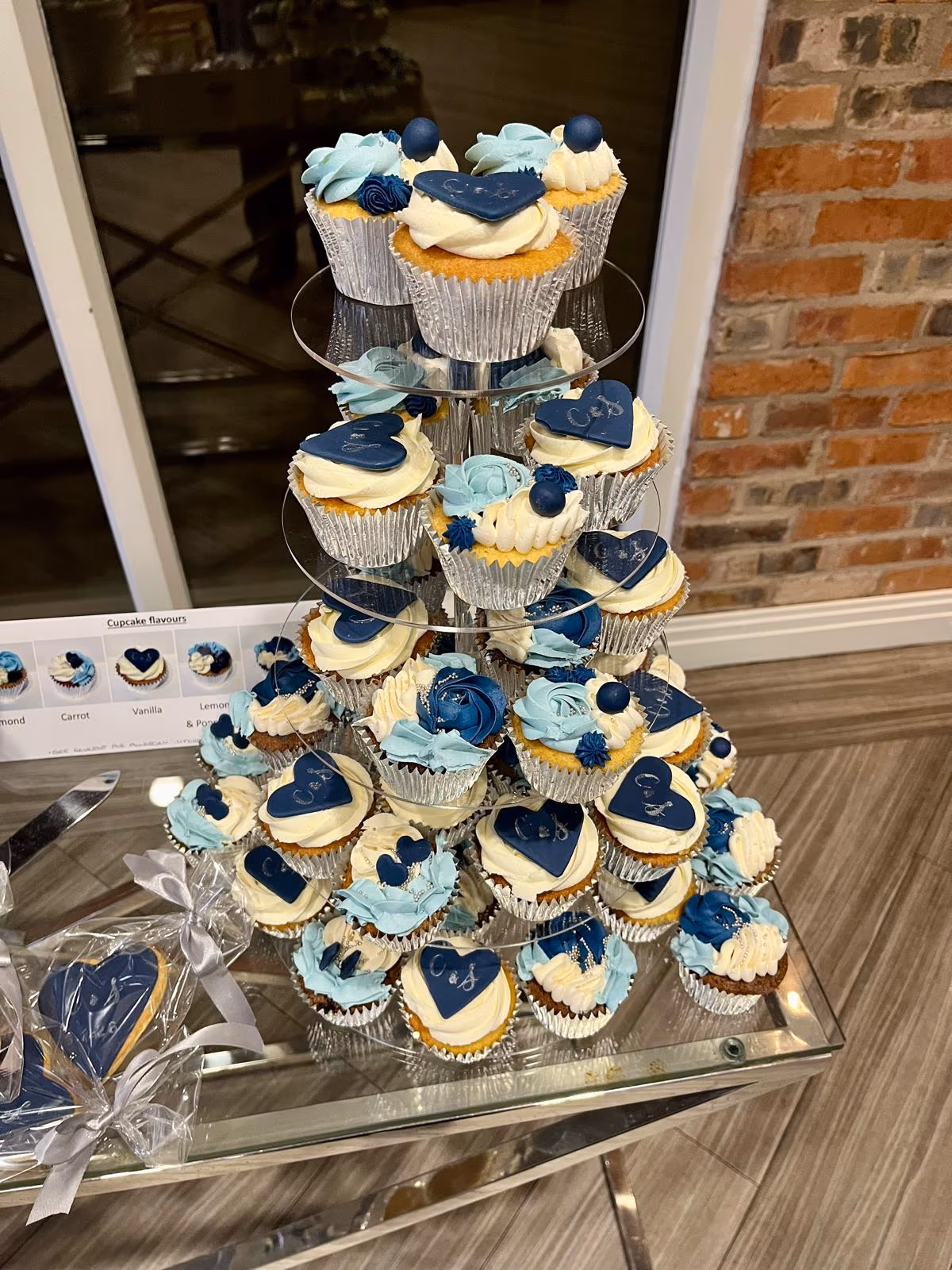 A multi-tiered display of wedding cupcakes decorated with white, light blue, and dark blue frosting, featuring heart-shaped toppers personalised with 'C & J', sugar flowers, and berries. Matching heart-shaped cookies are also visible on the table.