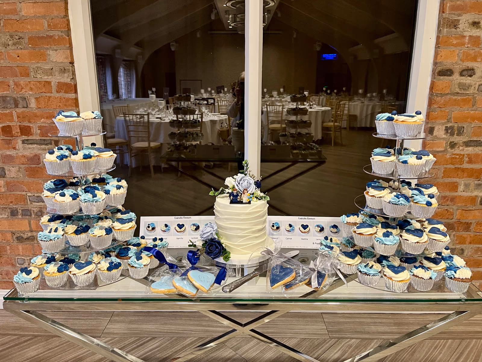 A wedding dessert display featuring a white tiered wedding cake with a bride and groom topper and blue floral accents, two multi-tiered cupcake stands with blue and white cupcakes, and individually wrapped blue heart-shaped cookies, all set on a mirrored table in a wedding reception venue.