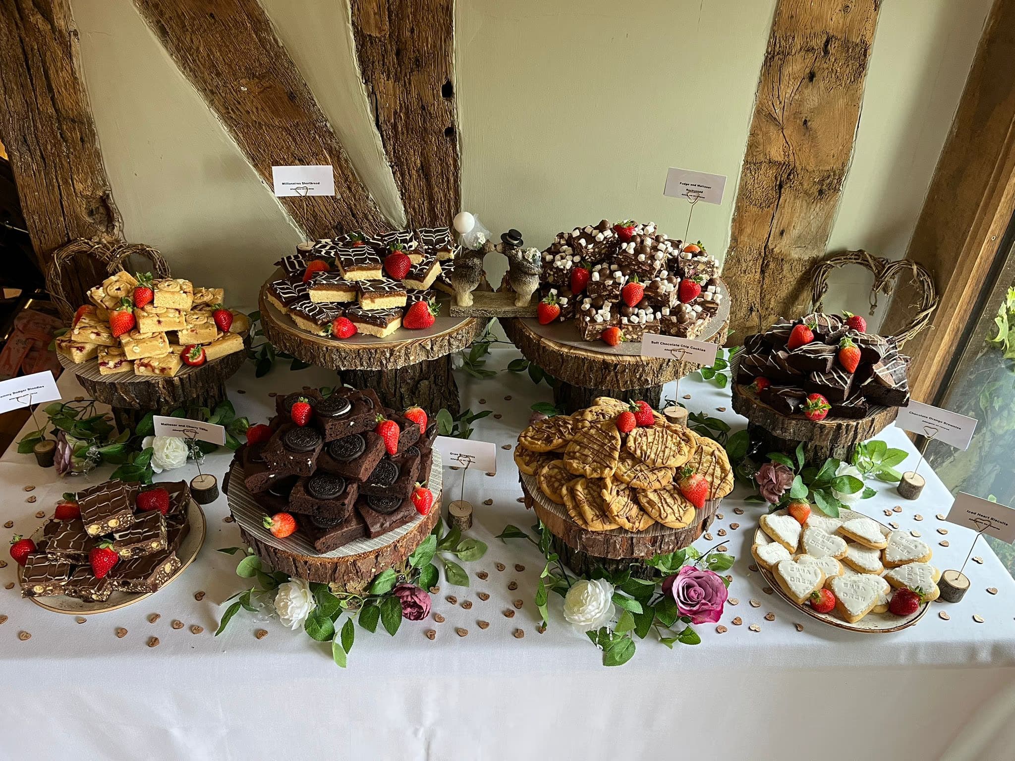 A rustic wedding dessert table laden with various baked goods including millionaire's shortbread, rocky road, Oreo brownies, chocolate chip cookies, and personalized iced heart biscuits, all garnished with fresh strawberries and floral decorations.