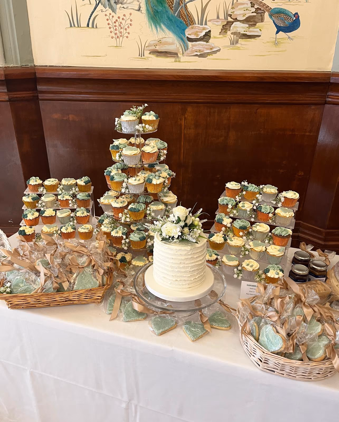 A wedding dessert table featuring a white textured cake topped with white and green flowers, surrounded by tiered stands of cupcakes in white, green, and gold wrappers, and baskets of individually wrapped heart-shaped cookies.