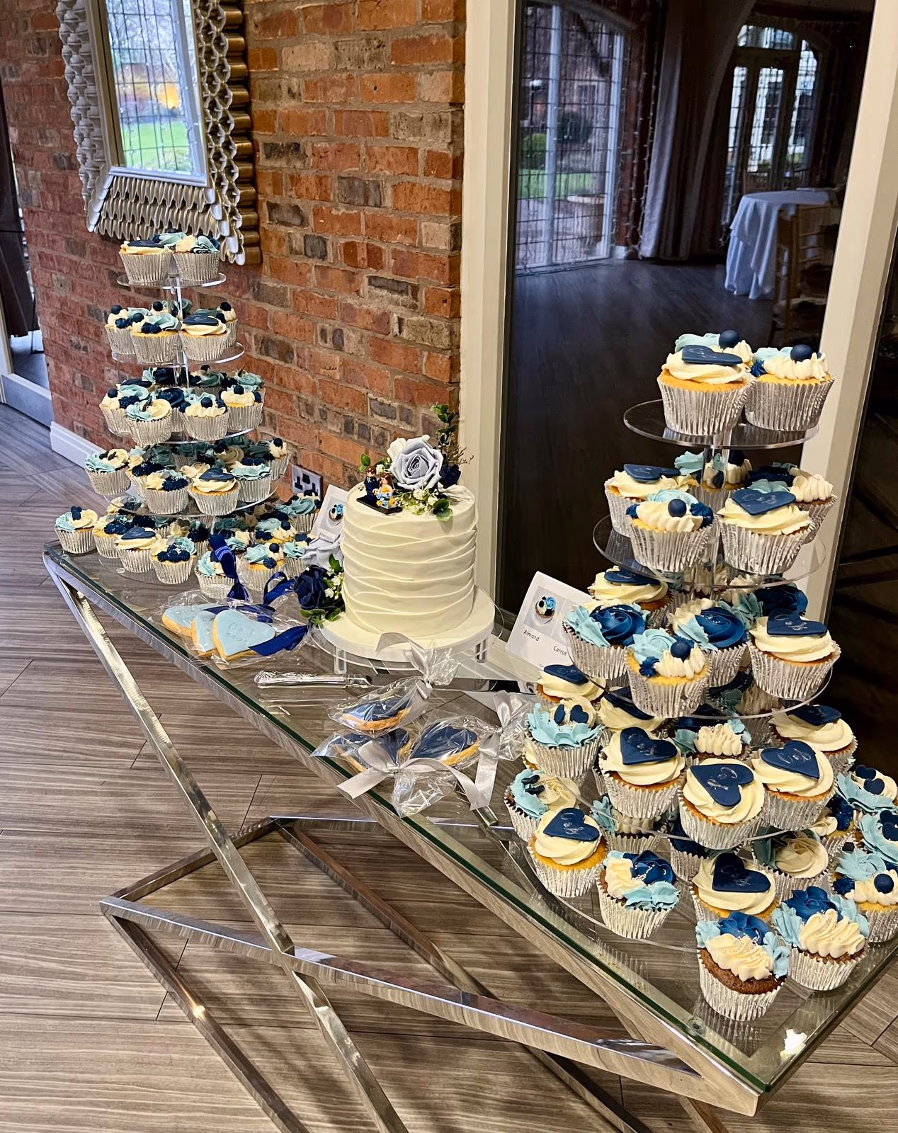 A dessert table featuring a white tiered cake decorated with blue and white flowers, surrounded by numerous cupcakes with blue and white frosting, and individually wrapped blue-themed cookies, all arranged on glass stands.