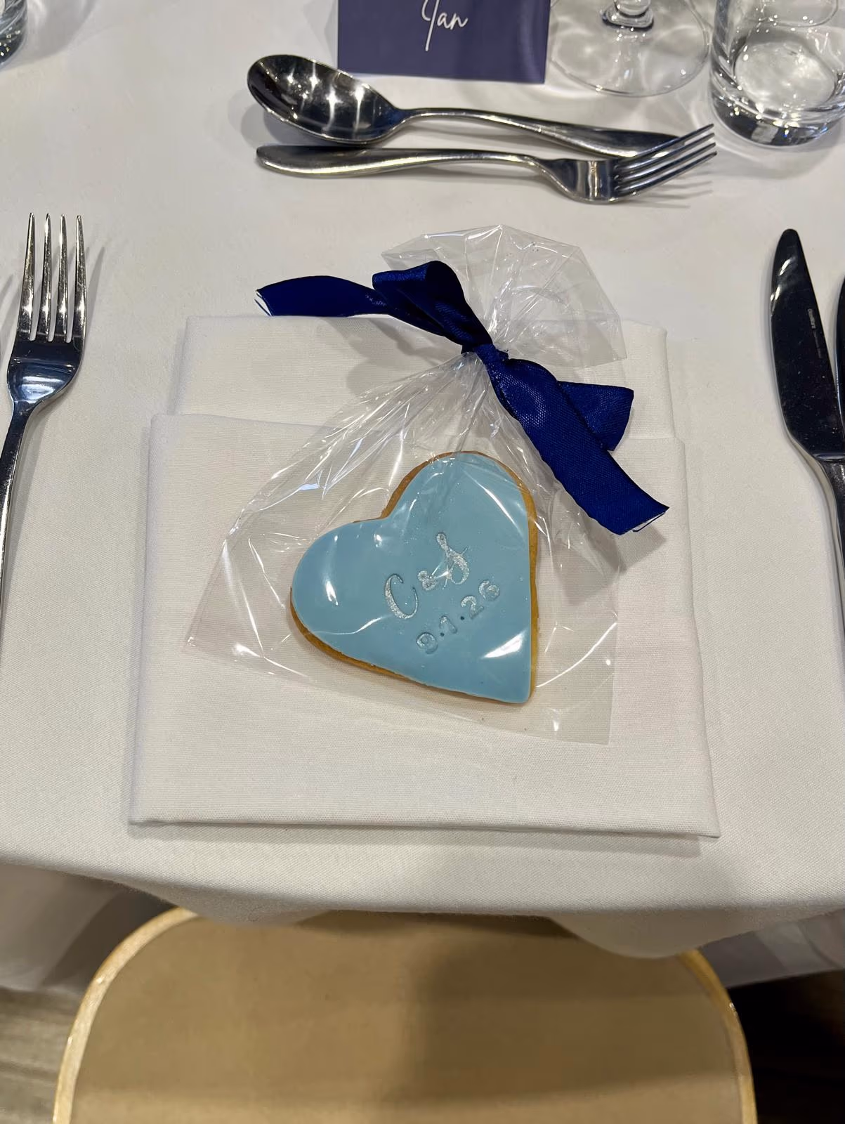 A light blue heart-shaped biscuit with white icing initials C & J and the date 9.1.26, packaged in a clear bag with a dark blue ribbon, set on a white napkin at a table.