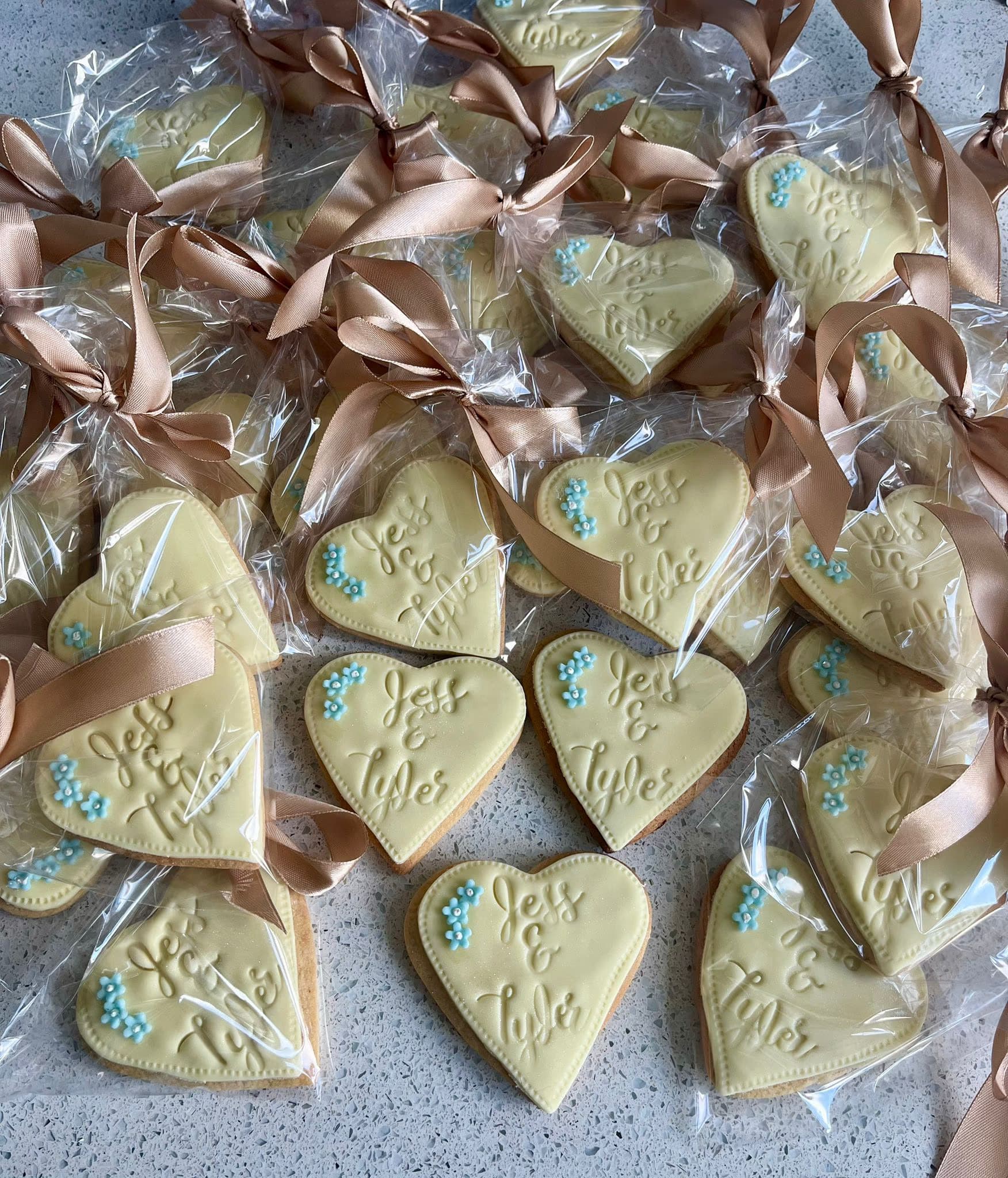A collection of individually wrapped heart-shaped cookies, each featuring cream icing with 'Jess & Tyler' embossed and small blue flower decorations, tied with a light brown ribbon.