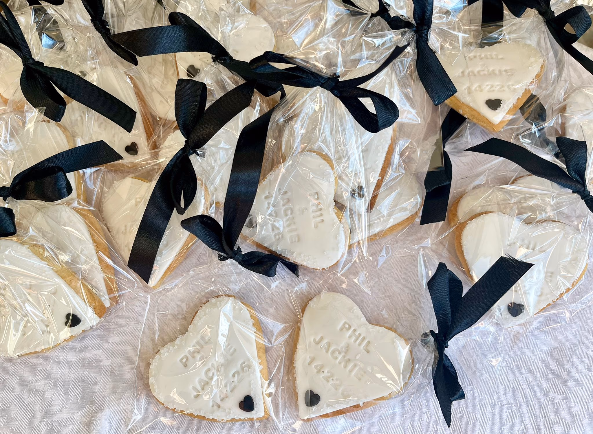 A close-up, top-down view of numerous heart-shaped cookies, each individually wrapped in clear plastic and tied with a black satin ribbon. The cookies have white icing with the embossed names 'PHIL JACKIE' and the date '14.2.26', along with a small black heart.