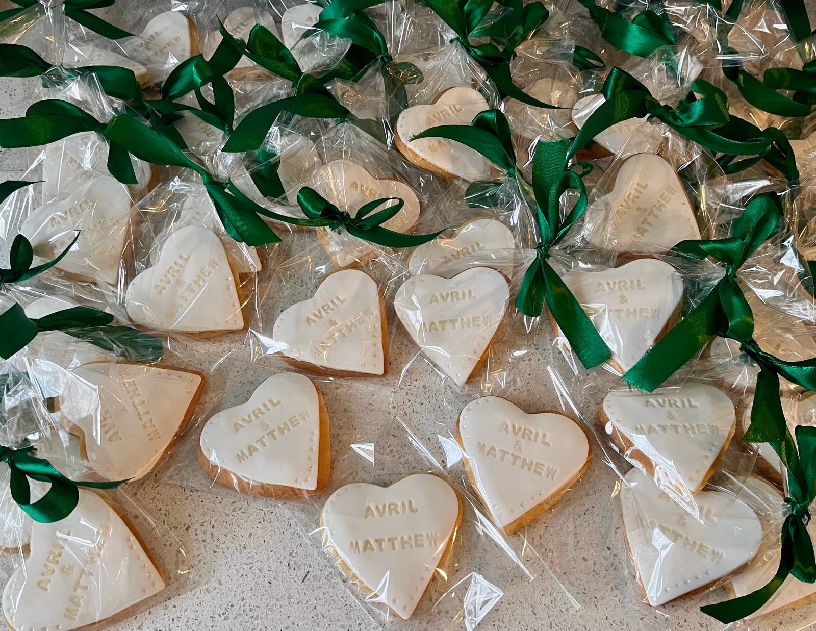 A close-up view of numerous heart-shaped cookies, each individually wrapped in clear plastic, decorated with white icing, the names 'AVRIL & MATTHEW' in gold, and tied with a vibrant green ribbon.