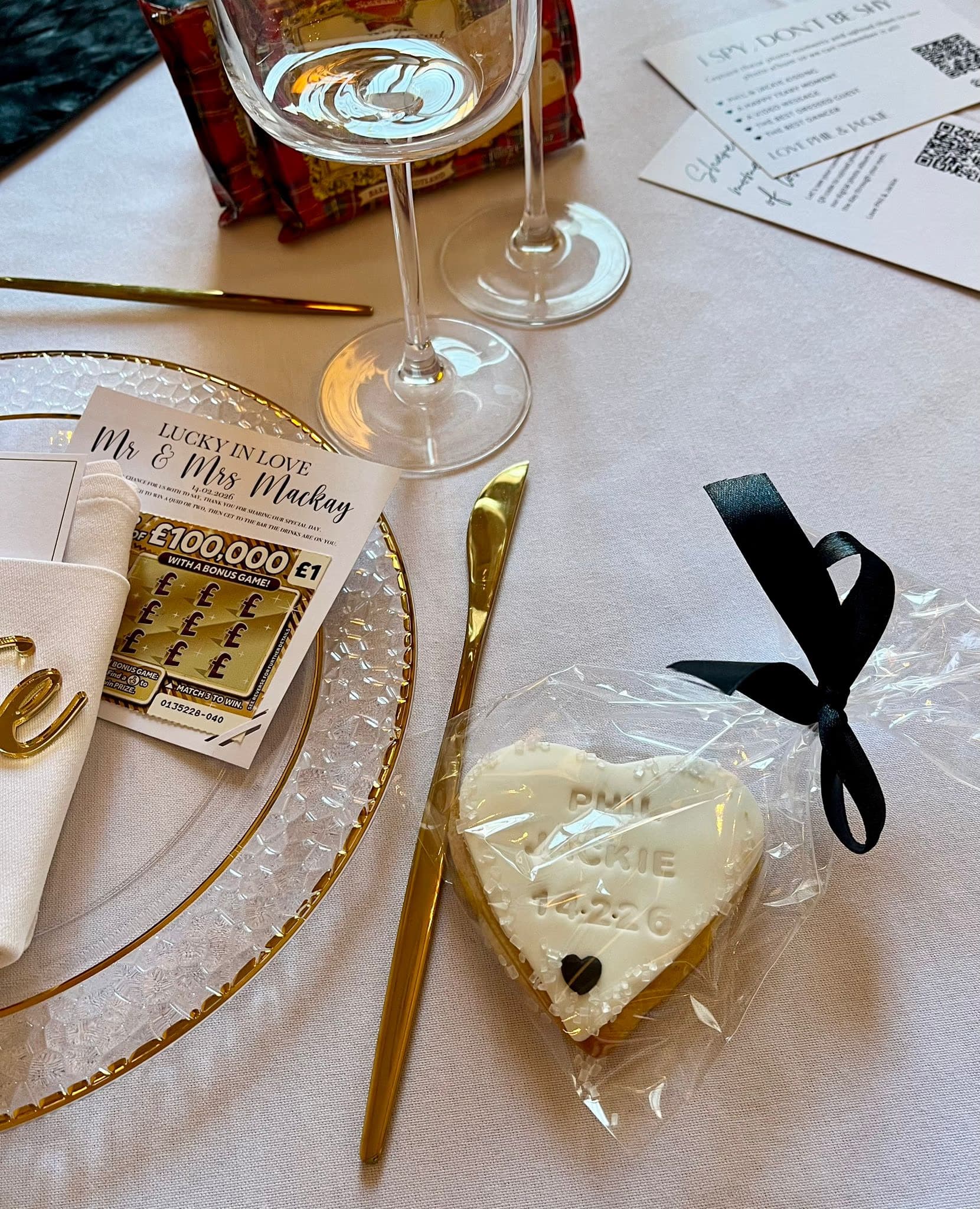 A white iced heart-shaped cookie, individually wrapped in clear plastic with a black ribbon, serving as a wedding favour with the names Phil and Jackie and the date 14.2.26 inscribed, placed on a table setting next to a 'Lucky in Love' scratch card.