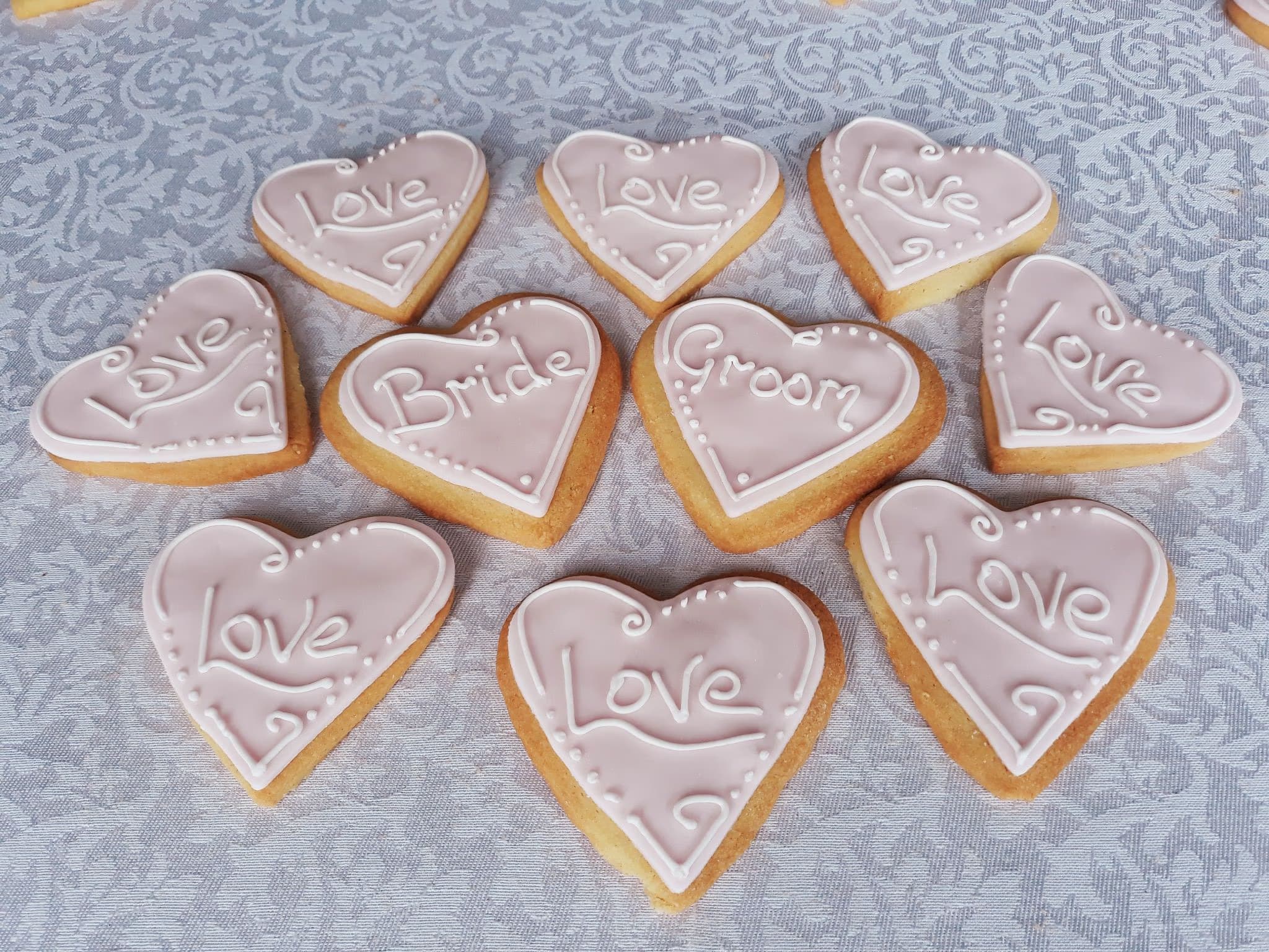 A collection of heart-shaped cookies with pink icing and white decorative text, including 'Love', 'Bride', and 'Groom', arranged on a patterned white tablecloth.