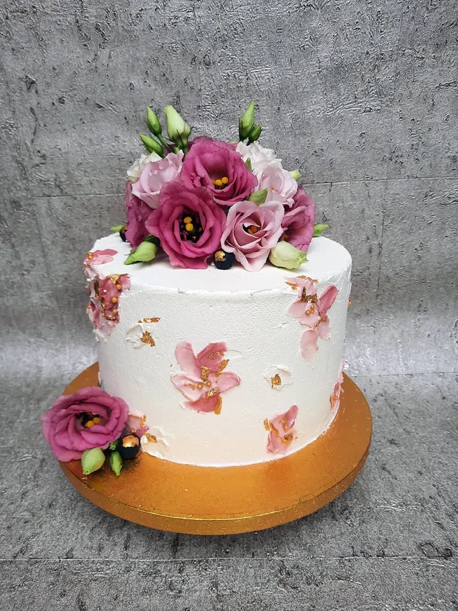 A single-tier white textured cake decorated with a vibrant bouquet of pink and magenta flowers, green buds, and dark berries on top, and delicate pink buttercream flowers with gold leaf accents on the sides. The cake rests on a gold cake board.