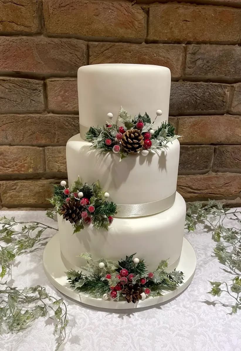 A three-tier white fondant cake decorated with frosted winter foliage, red berries, and pinecones, set against a rustic brick wall.