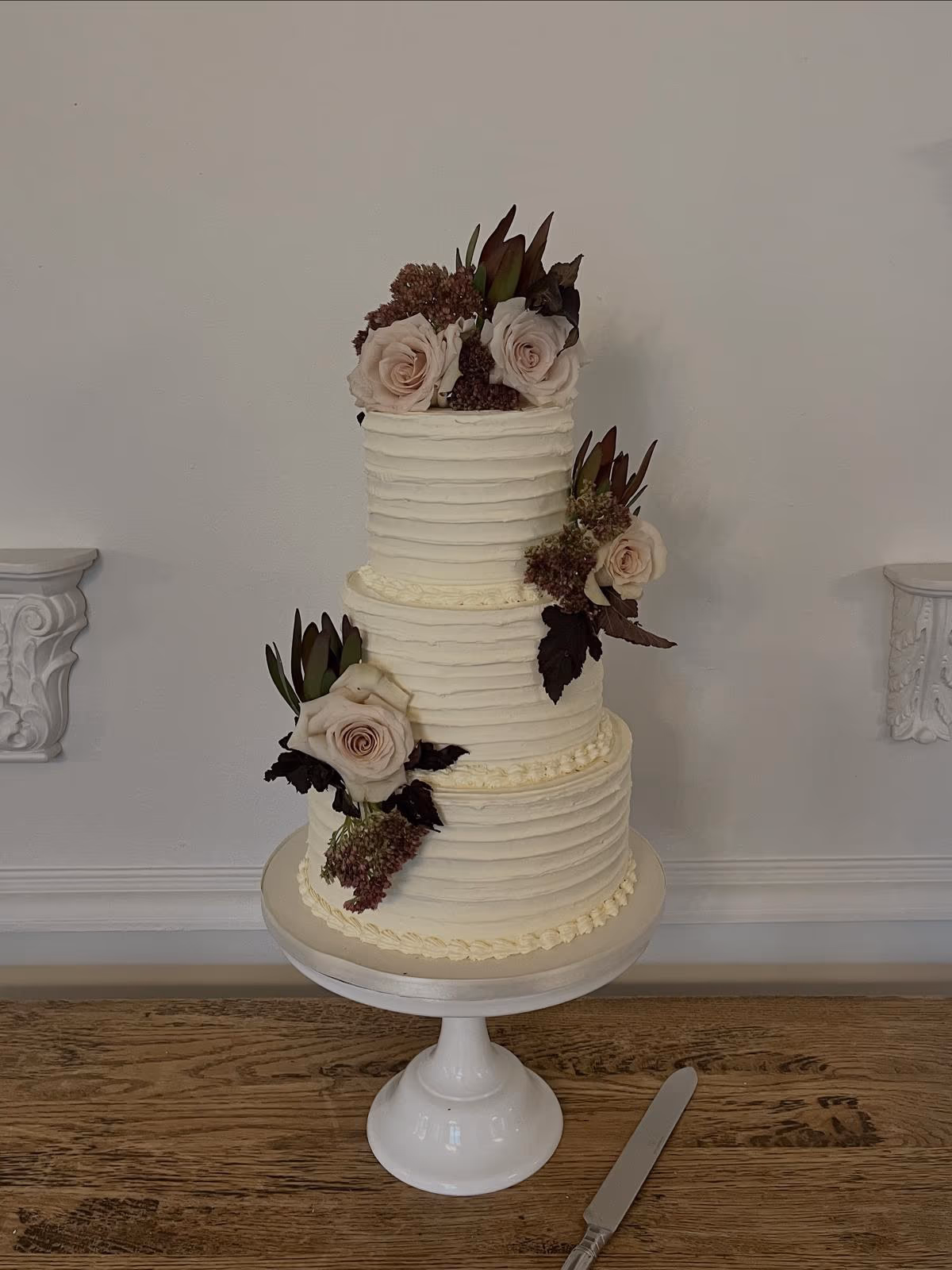 Three-tier cream textured buttercream wedding cake with blush roses and dark foliage