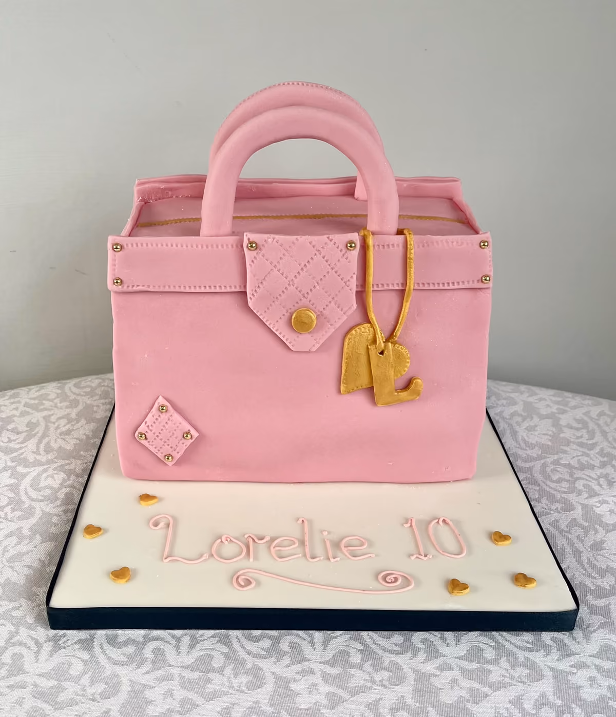 A 10th birthday cake for Lorelie shaped as a pink quilted handbag with gold studs and a gold initial charm, on a white board with pink lettering and gold hearts.