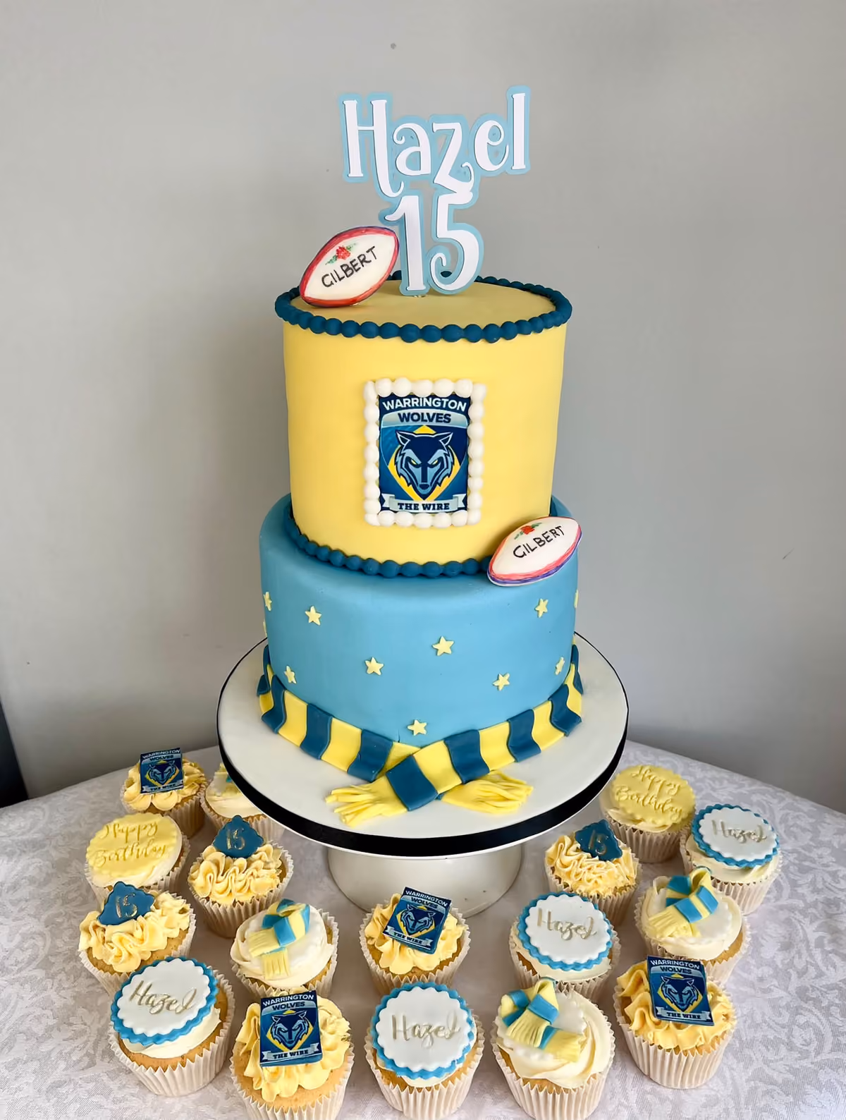 A two-tier 15th birthday cake for Hazel in Warrington Wolves yellow and blue, with the club badge, stars, a striped scarf and sugar rugby balls, topped with a 'Hazel 15' topper and surrounded by matching cupcakes.