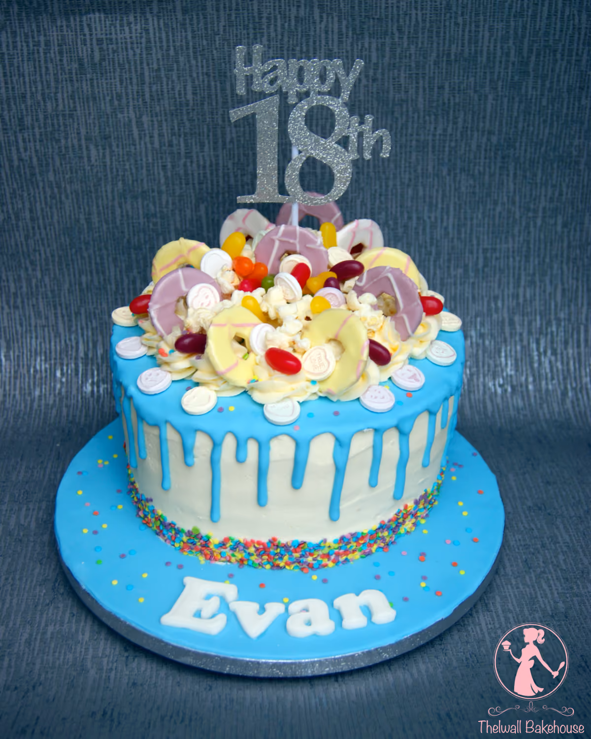 An 18th birthday cake with white frosting, blue drip icing, and a colorful assortment of candies, popcorn, and white discs on top. A silver glitter 'Happy 18th' topper is visible, and the blue cake board is adorned with sprinkles and the name 'Evan' in white.