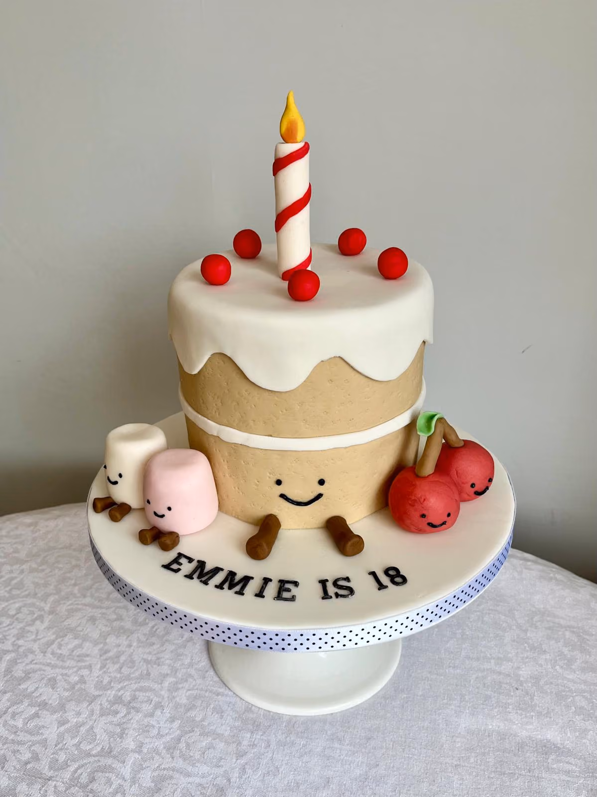 A fun 18th birthday cake for Emmie made to look like a smiling cartoon cake with white icing, a striped candle and red balls, joined by smiling marshmallows and cherries on a board reading 'Emmie is 18'.