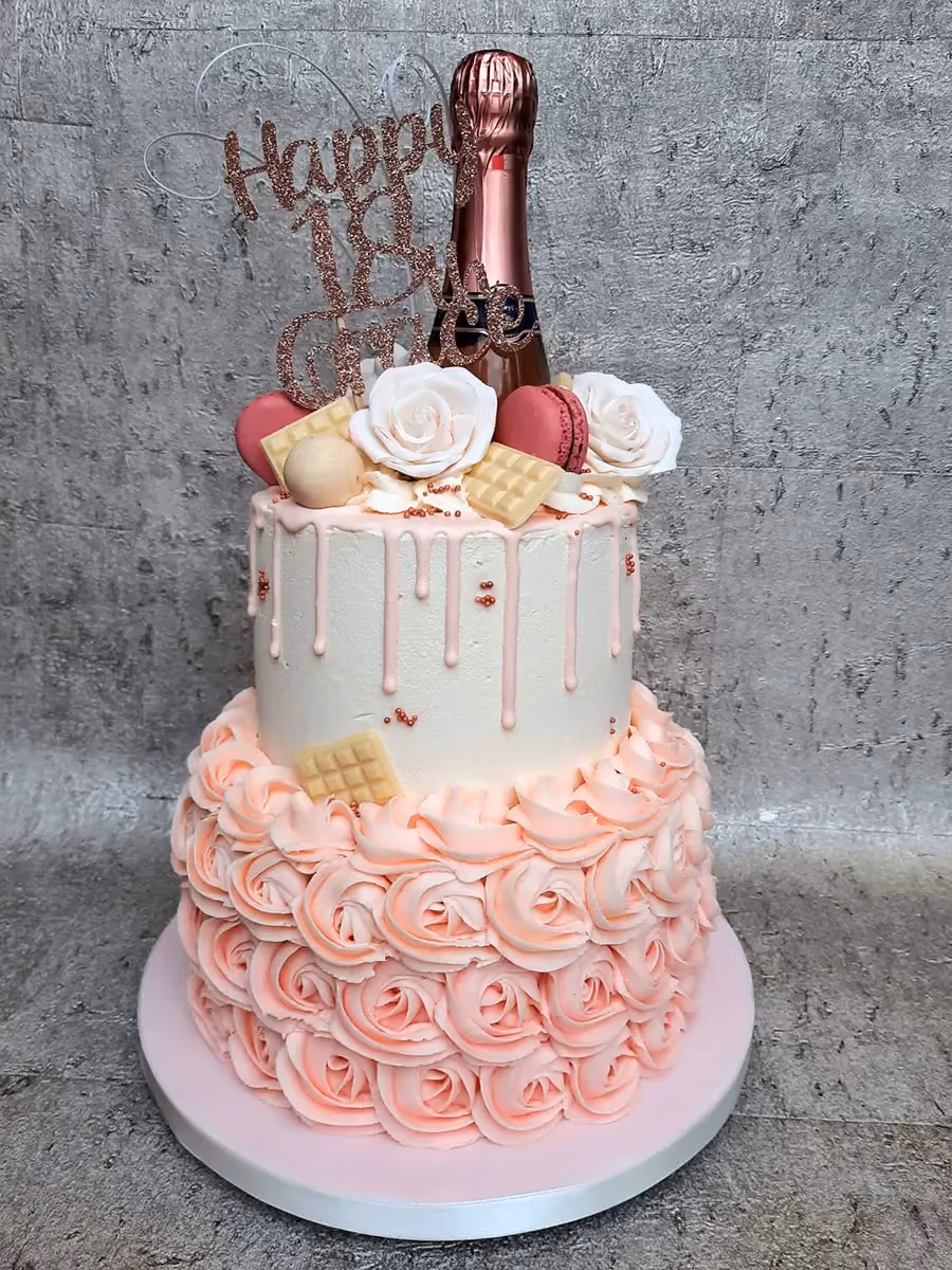 A two-tier 18th birthday cake featuring a white top tier with pink drip icing and sprinkles, and a pink bottom tier decorated with buttercream roses. The cake is adorned with a rose gold champagne bottle, a 'Happy 18th Birthday' topper, white sugar roses, pink macarons, and white chocolate bars.
