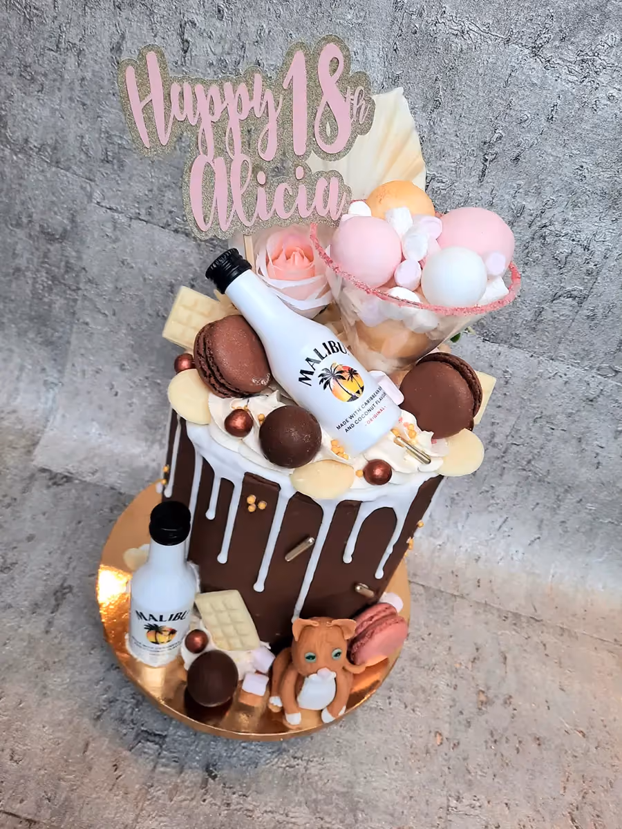 A lavish 18th birthday chocolate drip cake topped with a 'Happy 18th Alicia' sign, adorned with miniature Malibu bottles, chocolate macarons, white chocolate pieces, a cup filled with pink and white sweets and marshmallows, a pink rose, and a small cat figurine.