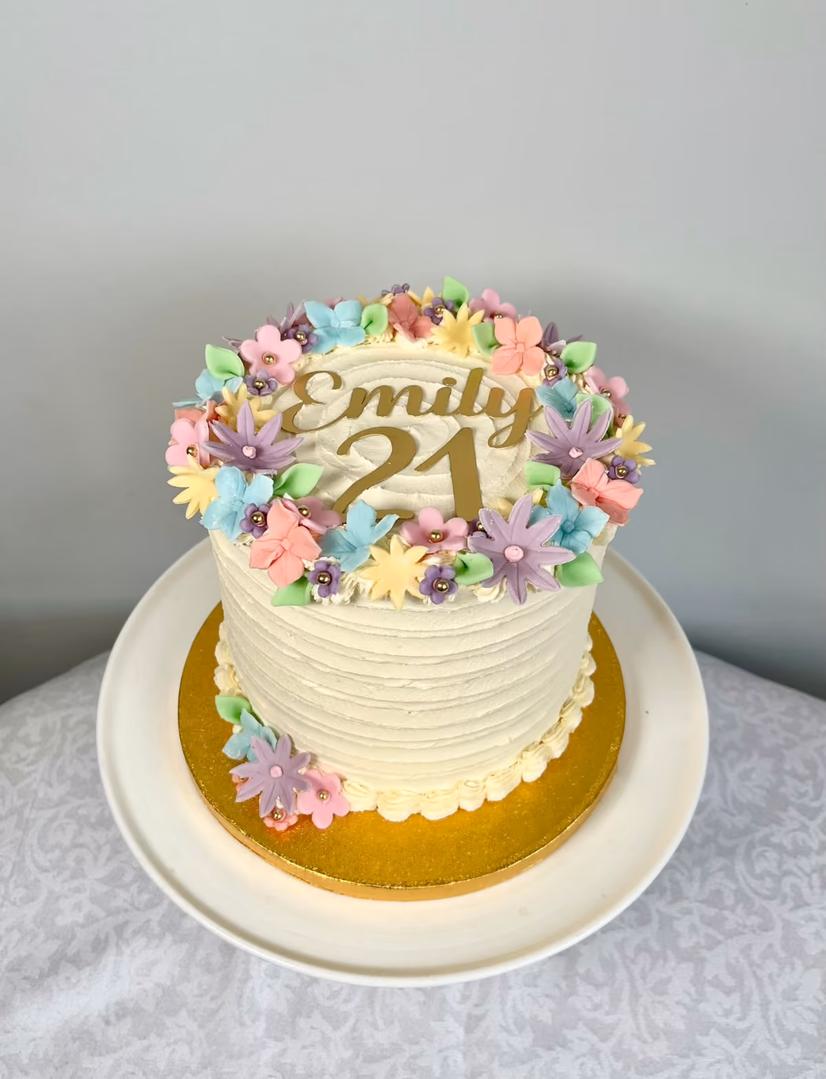 A textured ivory buttercream 21st birthday cake for Emily, topped with a gold 'Emily 21' topper inside a ring of pastel sugar flowers, on a gold board.