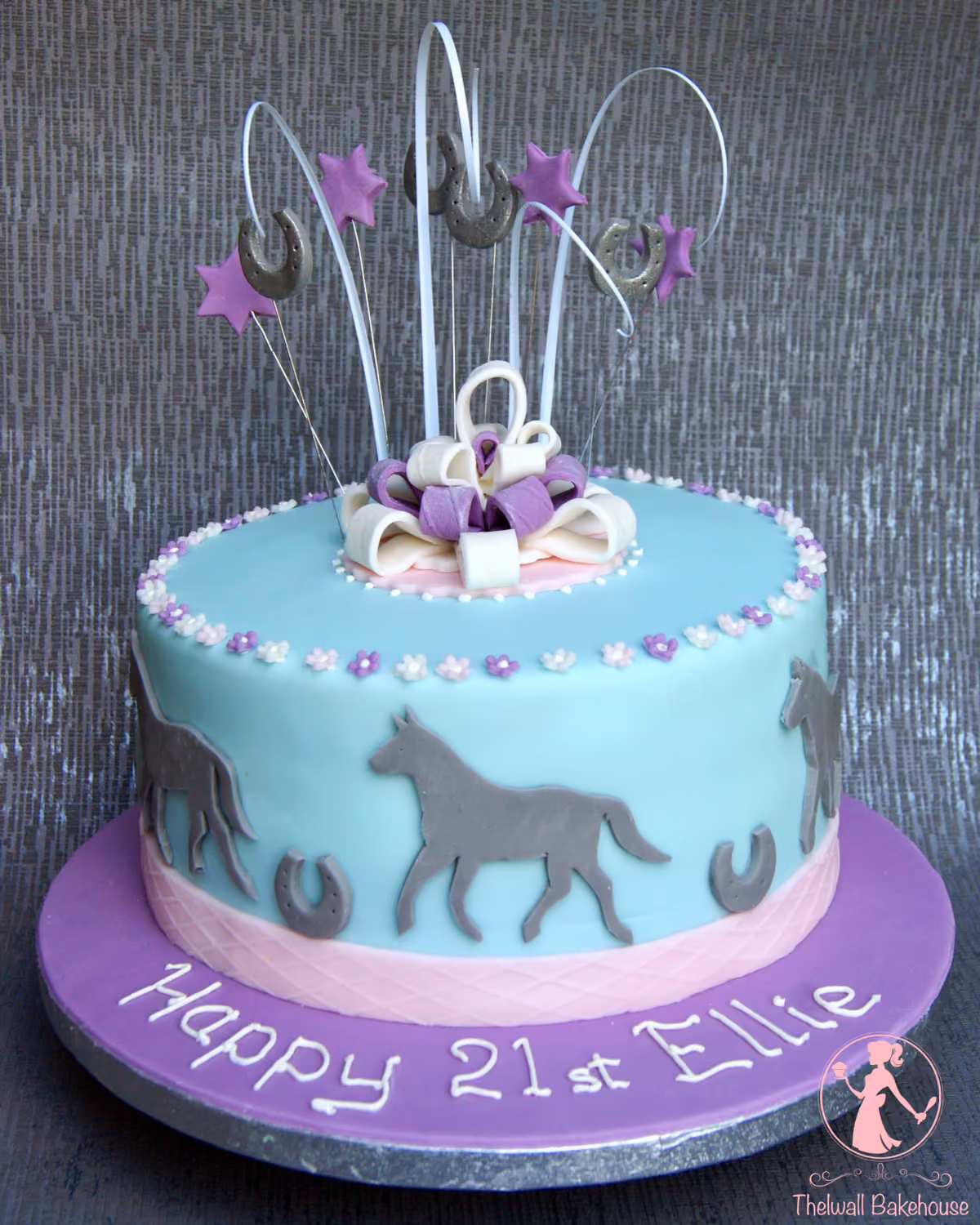 A light blue round 21st birthday cake decorated with grey horse silhouettes and horseshoes around the side, topped with a white and purple ribbon bow, and wire toppers featuring purple stars and grey horseshoes. The purple cake board is inscribed with 'Happy 21st Ellie'.