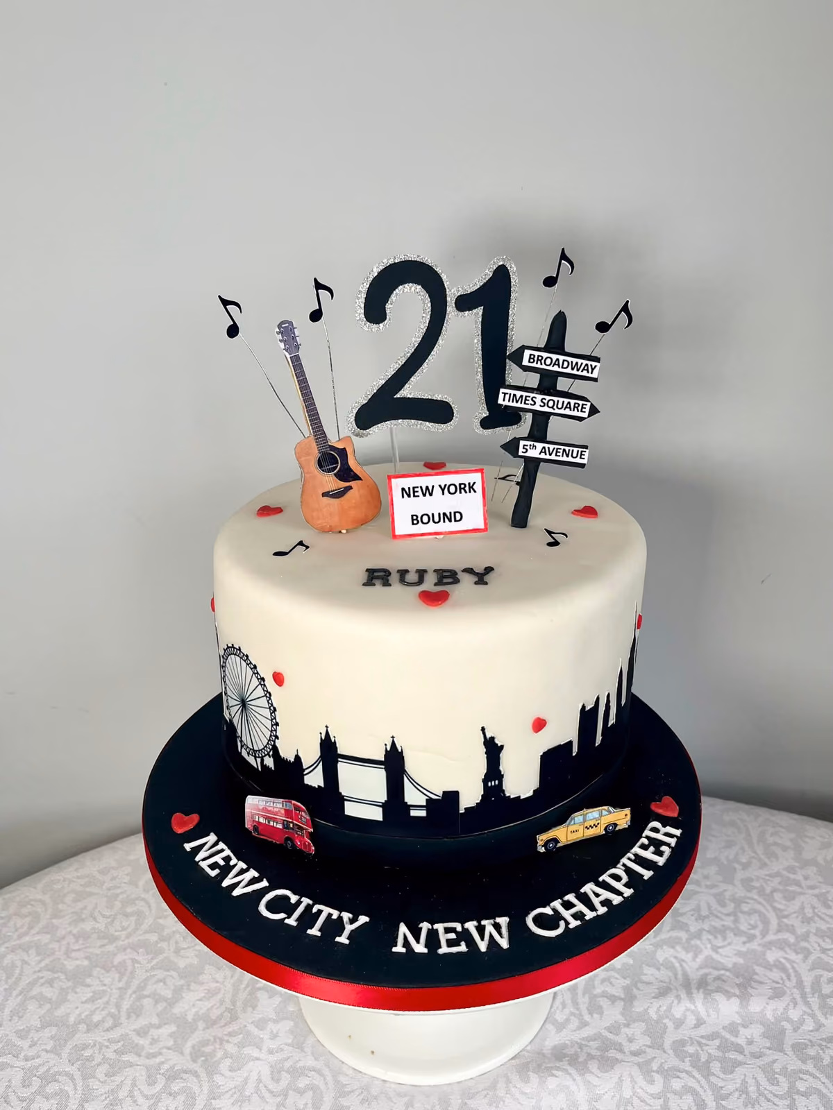 A 21st birthday cake for Ruby with a black London and New York skyline, a guitar, music notes, Broadway and Times Square street signs and a 'New York Bound' sign, on a board reading 'New City New Chapter'.