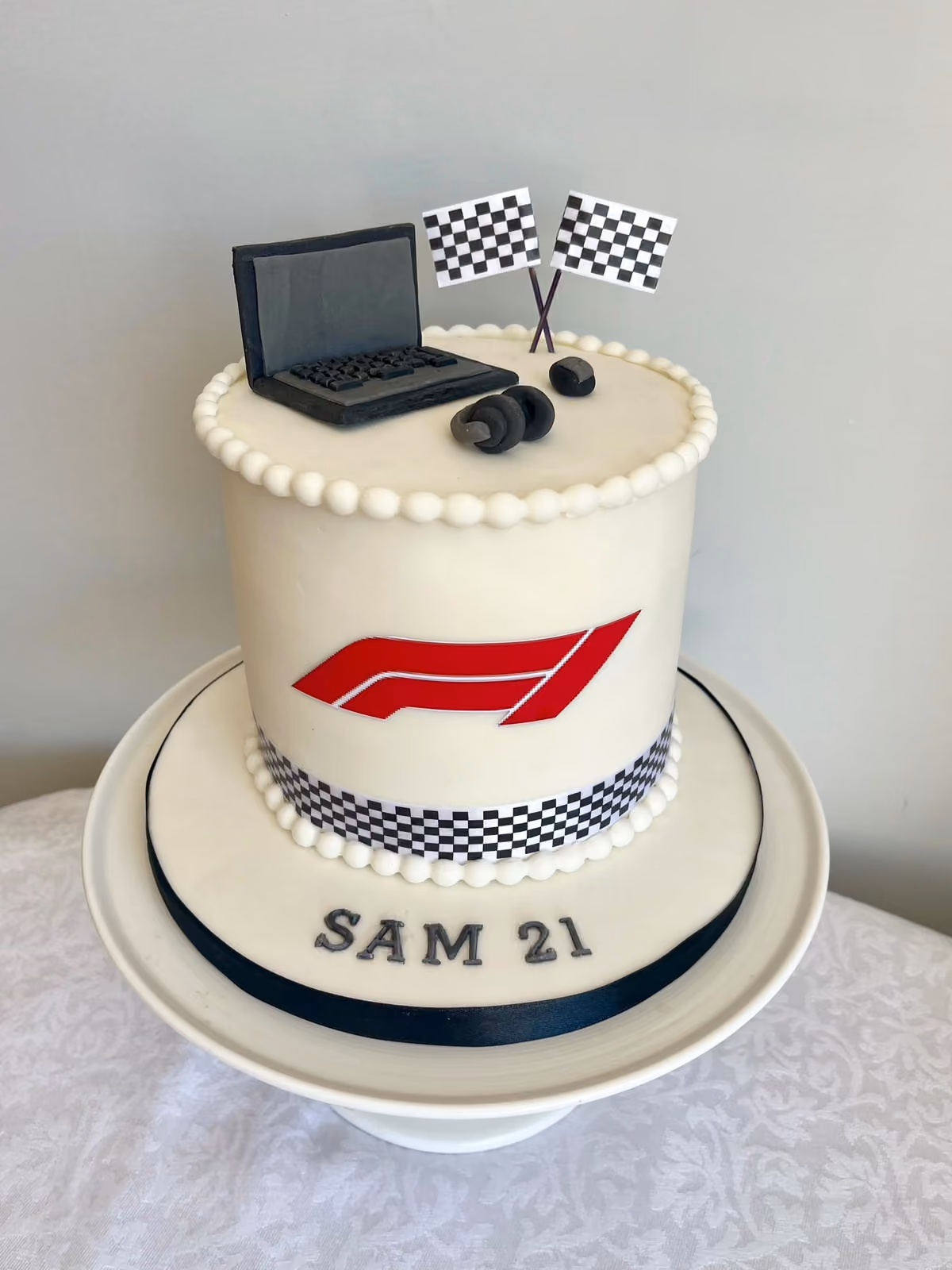 A white Formula 1 themed 21st birthday cake for Sam with a red F1 logo and chequered band, topped with a sugar laptop, headphones and crossed chequered flags.