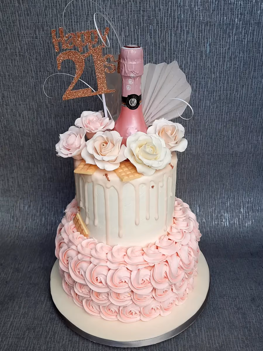 A two-tier 21st birthday cake with a pink rosette bottom tier and a white drip top tier, topped with a mini pink champagne bottle, pink and white sugar roses, and a glittery 'Happy 21st' topper.