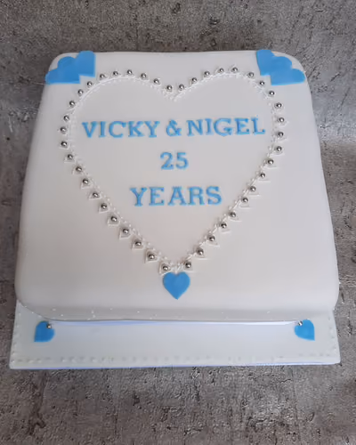 A white square anniversary cake for Vicky and Nigel, celebrating 25 years, decorated with blue text, a silver pearl heart border, and blue heart accents on the cake and board.