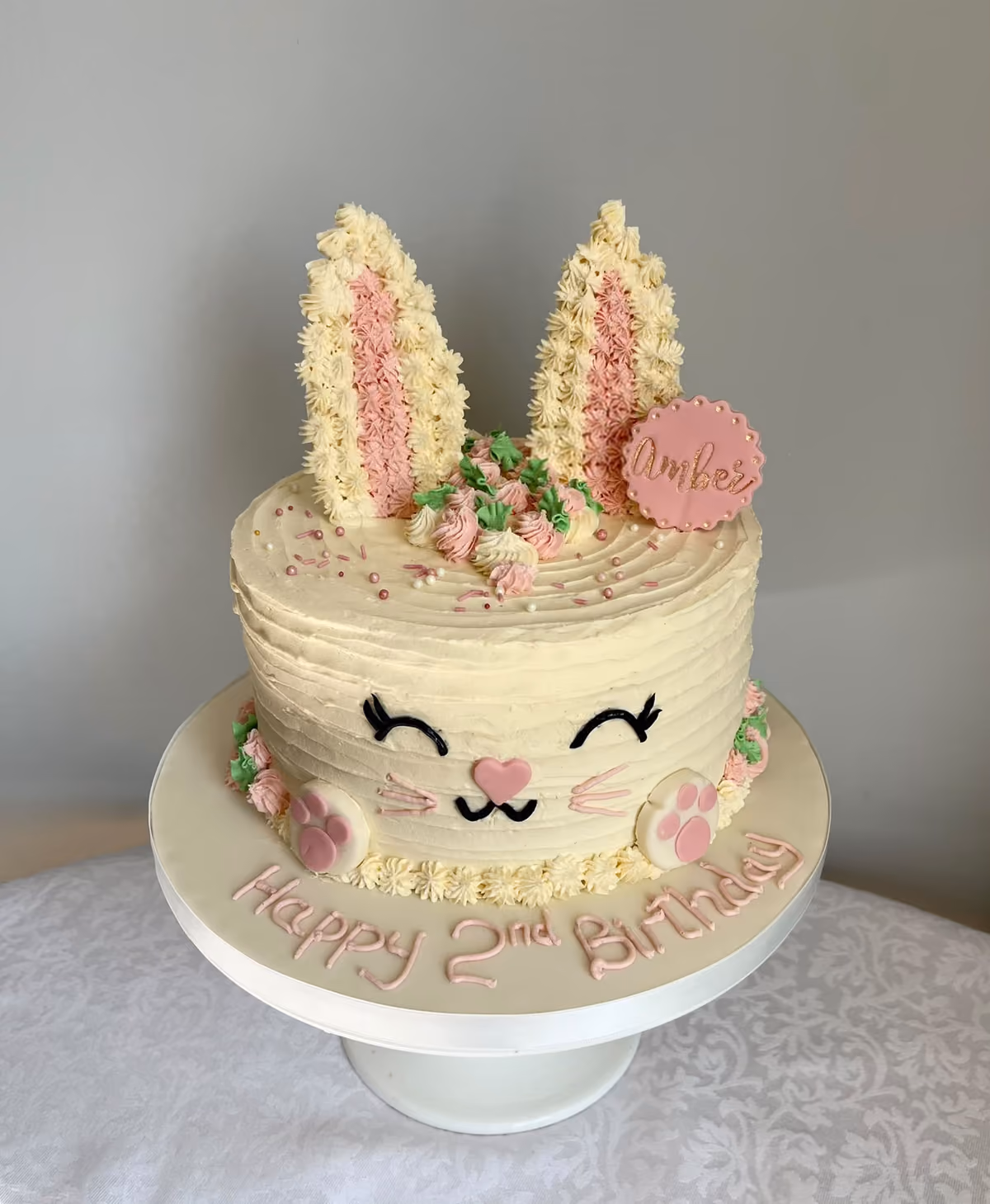 A bunny face 2nd birthday cake in cream buttercream with piped pink ears, a smiling face, piped pink flowers and 'Happy 2nd Birthday' on the board.