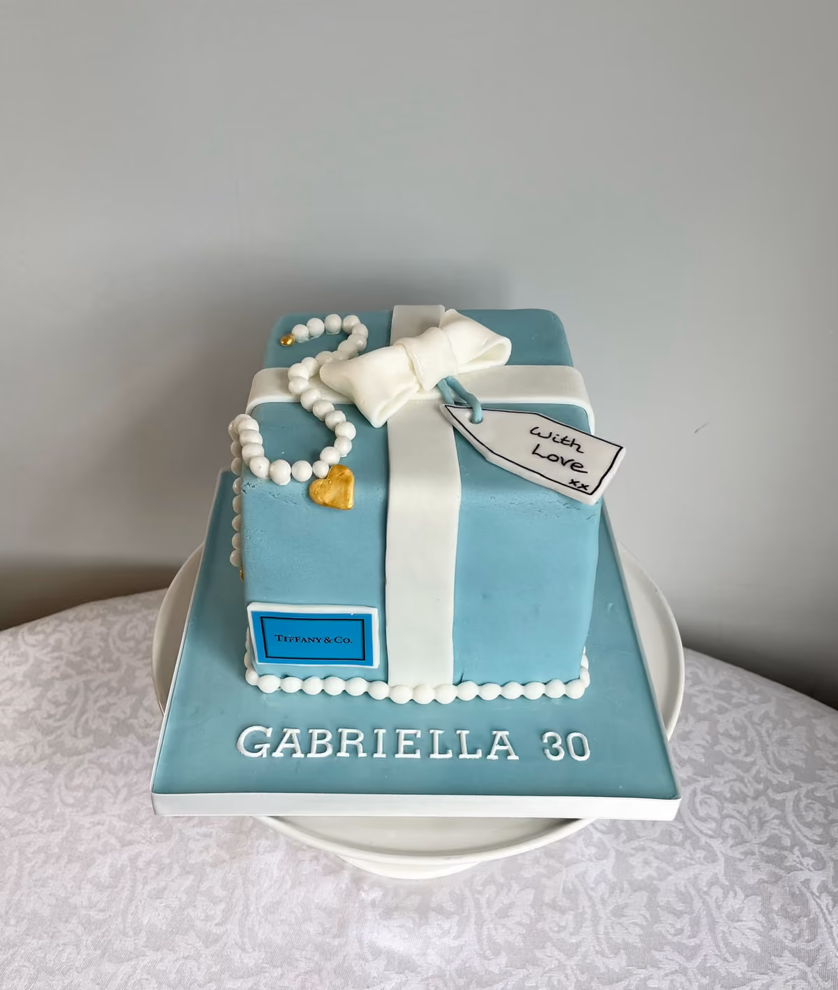 A 30th birthday cake for Gabriella shaped as a duck-egg blue gift box with a white ribbon and bow, a string of sugar pearls and a 'With Love' gift tag.