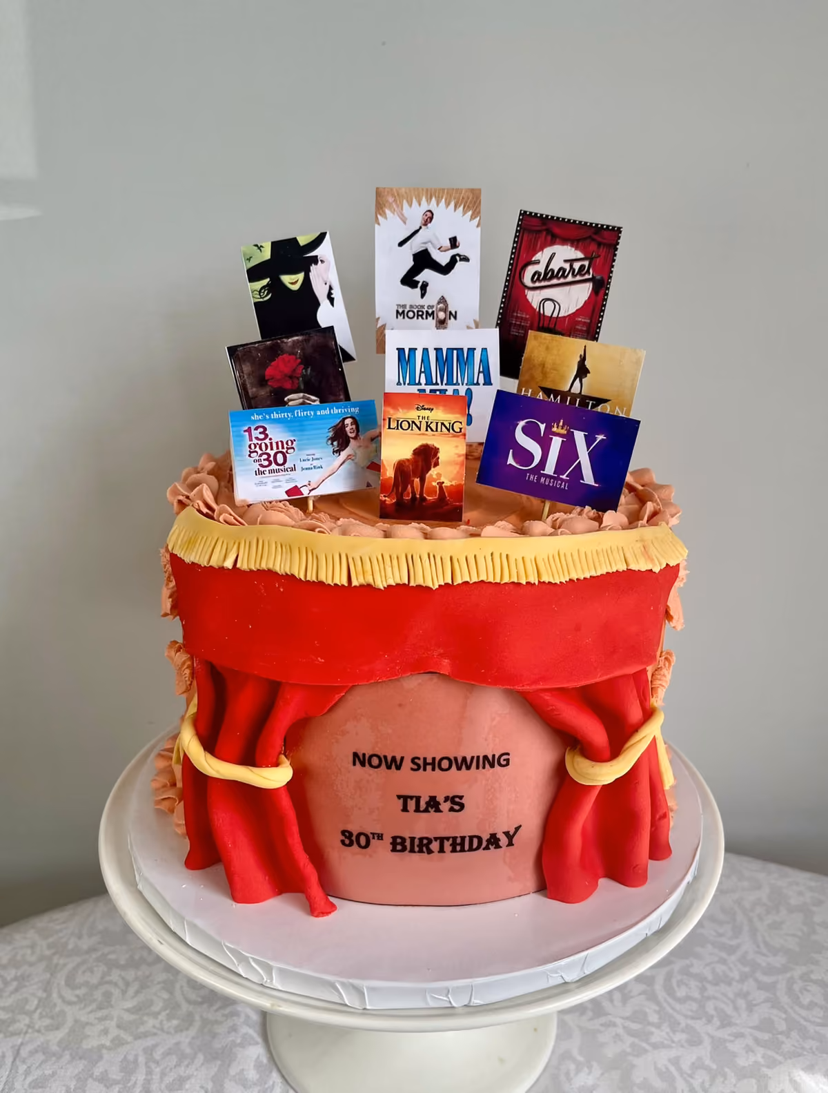 A musical theatre 30th birthday cake for Tia with red stage curtains and a 'Now Showing Tia's 30th Birthday' sign, topped with posters from shows including Six, Mamma Mia, The Lion King and Cabaret.