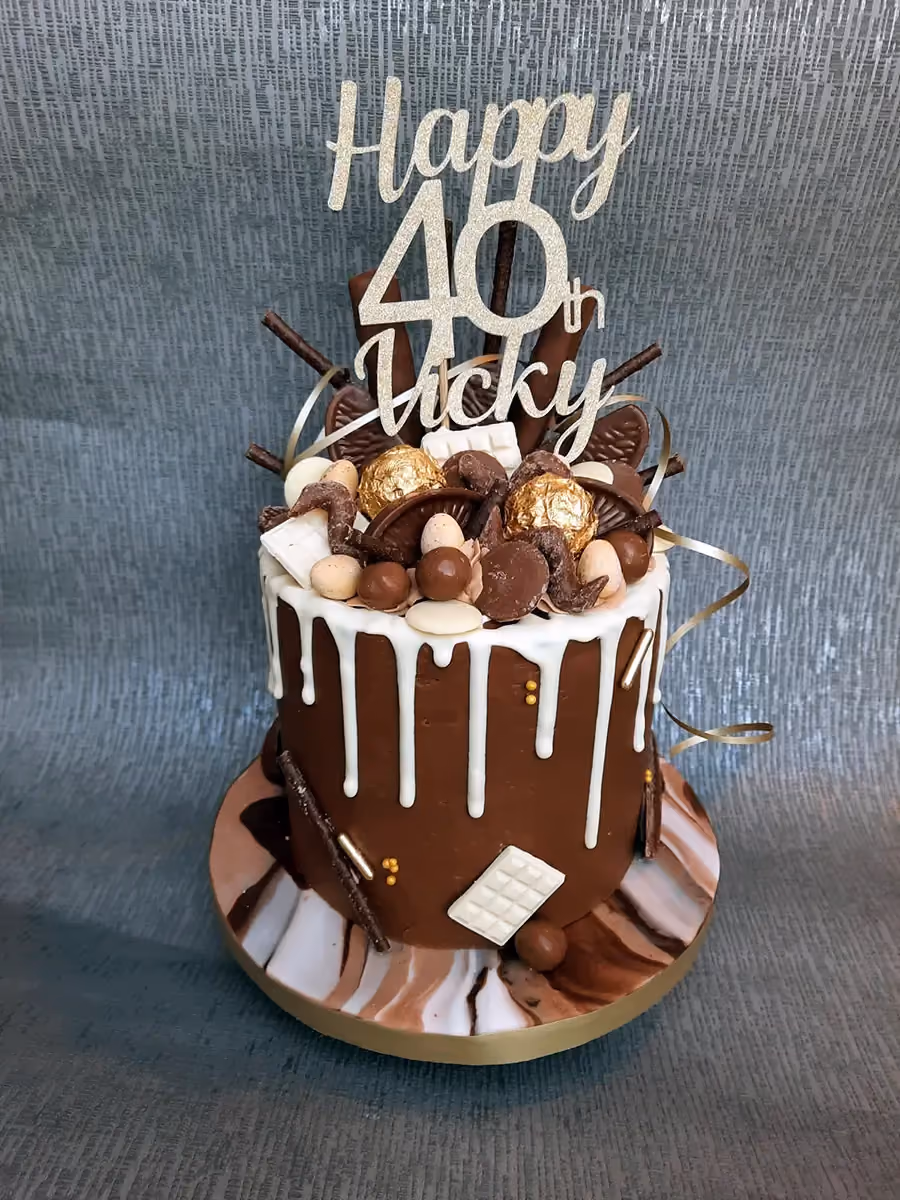 A tall chocolate drip cake with white chocolate drips, topped with an assortment of chocolates, gold foil candies, chocolate sticks, and a gold glitter "Happy 40th Vicky" cake topper. The cake sits on a marble-effect base with a gold rim.