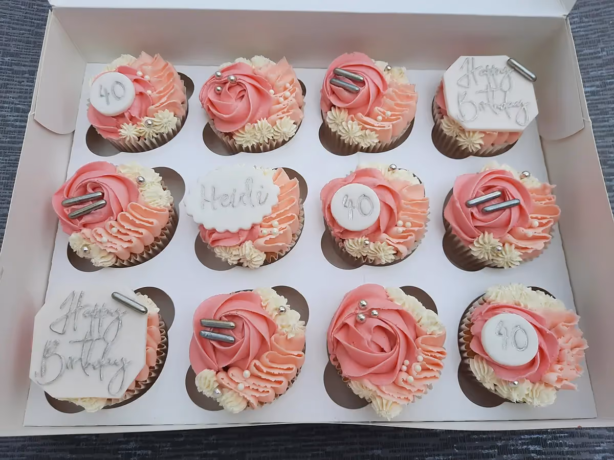 A dozen cupcakes in a white box, decorated with pink and cream frosting. Some cupcakes feature white sugar paste toppers with '40', 'Heidi', or 'Happy Birthday' written in silver, along with silver sprinkles.
