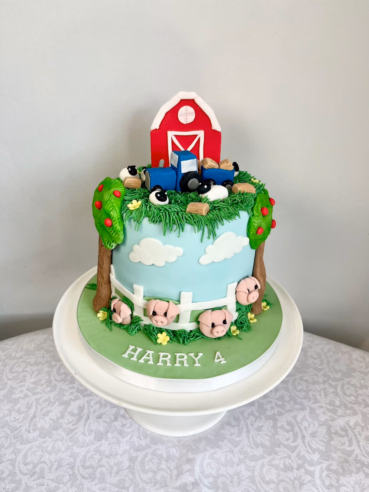 A farm themed 4th birthday cake for Harry with a red barn, a blue tractor, sheep and hay bales on a grassy top, and pigs, apple trees and a white fence around a sky-blue side.
