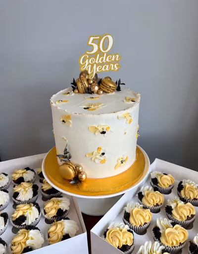 A white anniversary cake decorated with gold macarons, spheres, and black leaves, topped with a '50 Golden Years' topper, surrounded by matching gold, black, and white cupcakes.