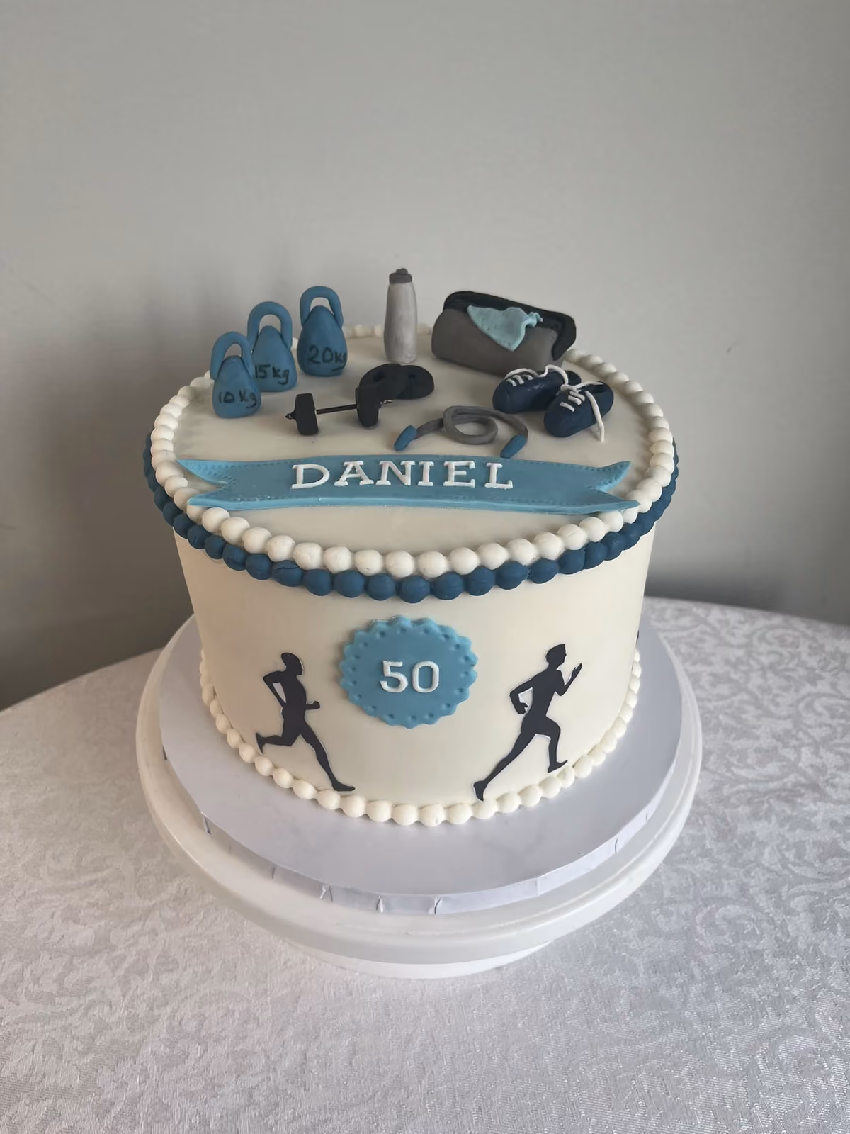 A white 50th birthday cake for Daniel with navy piped borders, running silhouettes on the side and sugar gym equipment on top, including kettlebells, a barbell, a water bottle, trainers and a kit bag.