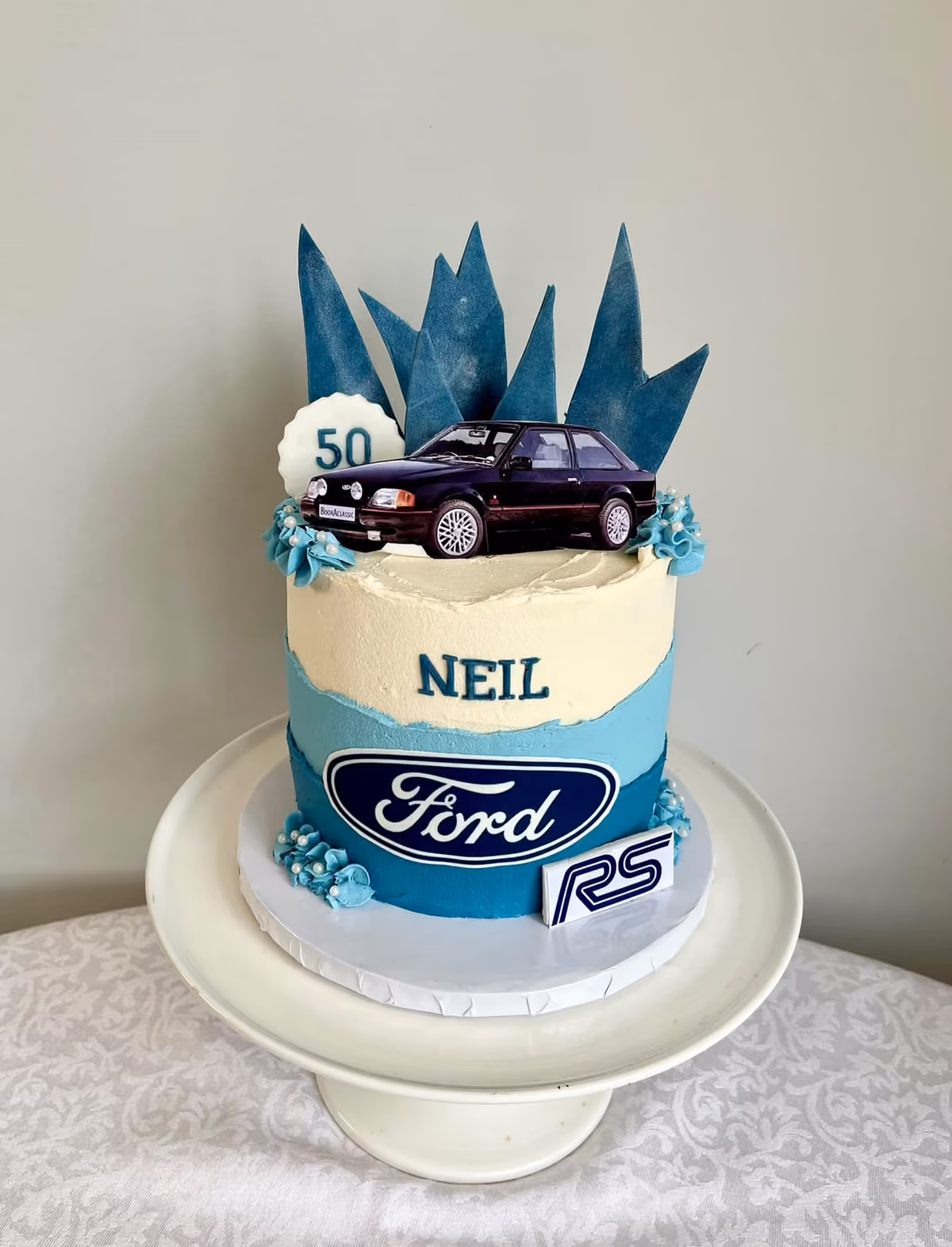 A blue and ivory 50th birthday cake for Neil with a Ford badge and RS logo, topped with a model Ford Escort RS, a '50' disc and dark blue shards.