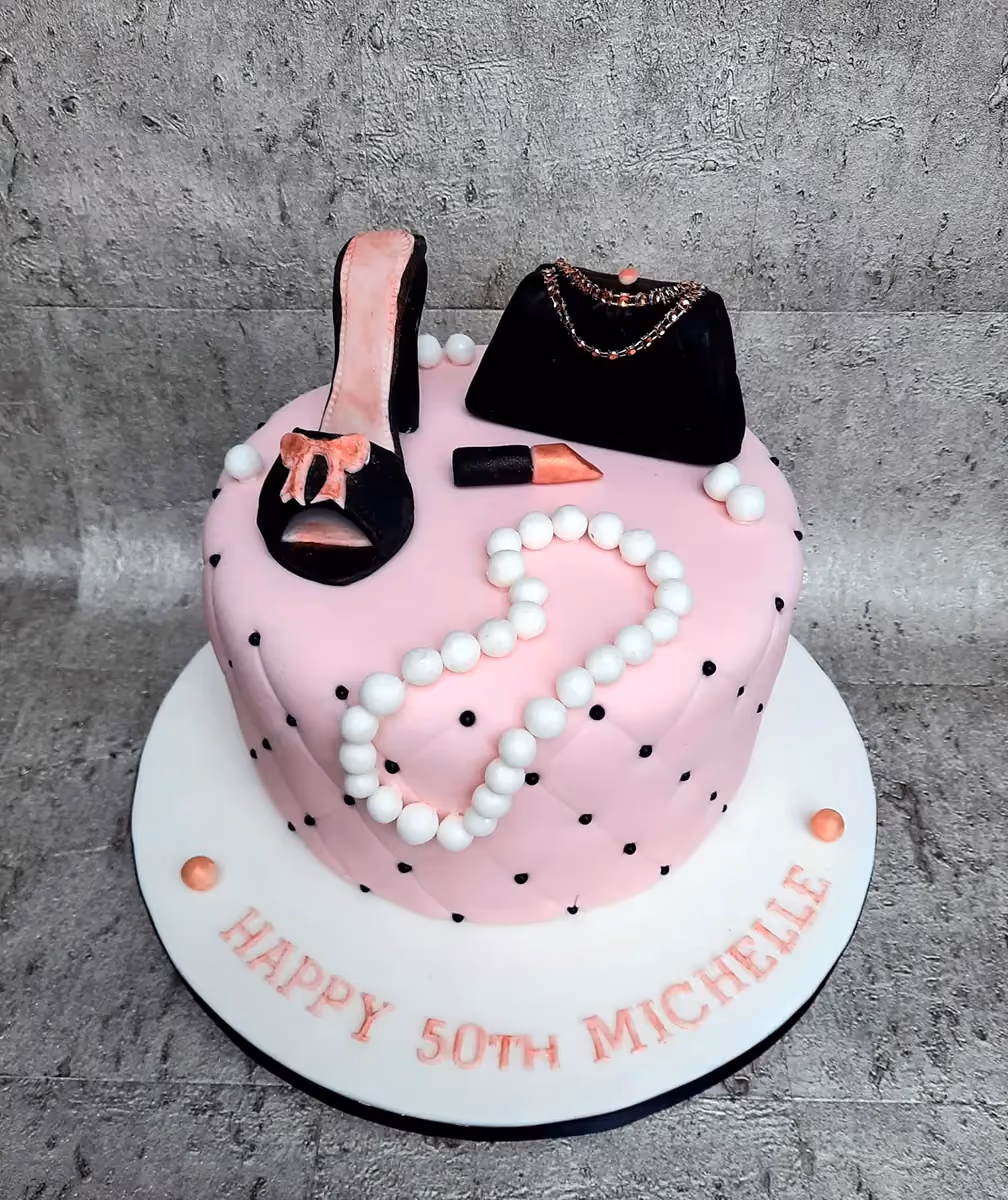 A pink quilted birthday cake decorated with a black high heel shoe, a black handbag, a red lipstick, and a white pearl necklace, with black polka dots on the cake sides. The white cake board has the inscription 'HAPPY 50TH MICHELLE' in pink letters.