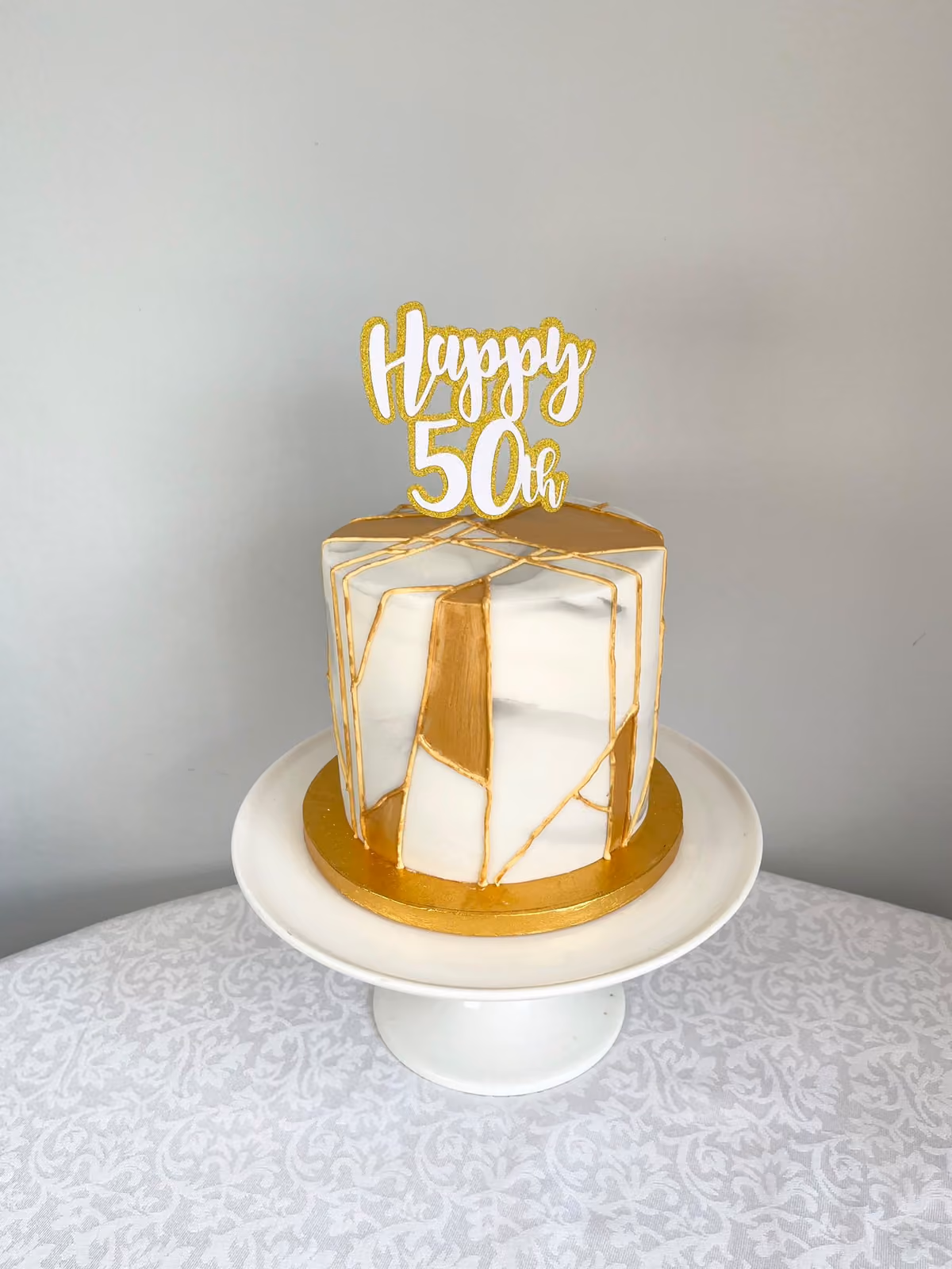 A white marble effect 50th birthday cake with gold kintsugi-style lines and panels, a gold board and a gold glitter 'Happy 50th' topper.
