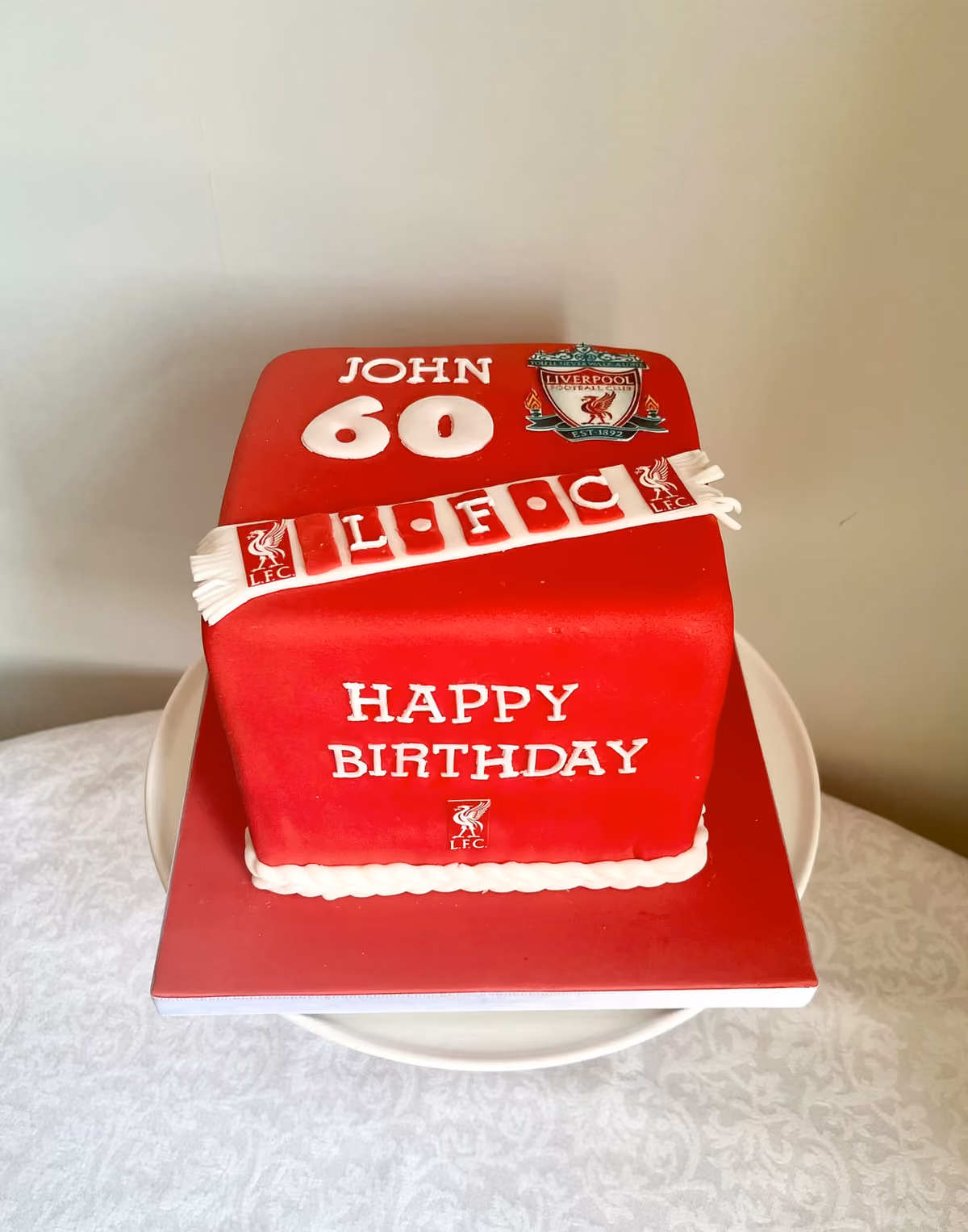 A square red Liverpool FC 60th birthday cake for John, with the club crest, a red and white 'LFC' scarf and 'Happy Birthday' in white lettering.