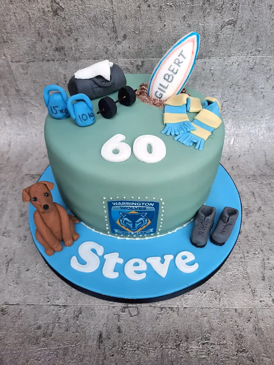 A round 60th birthday cake for Steve, featuring a pale green main tier decorated with gym equipment (kettlebells, barbell, gym bag), a rugby ball, a blue and yellow scarf, and the number '60'. The cake sits on a light blue base adorned with a brown dog, two grey hiking boots, and the name 'Steve' in white letters, along with a 'Warrington Wolves' logo on the side of the green tier.