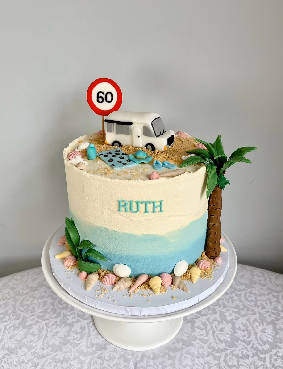 A beach themed 60th birthday cake for Ruth with blue ombre buttercream, a sugar campervan and '60' sign on a sandy top, a palm tree, and seashells around the base.
