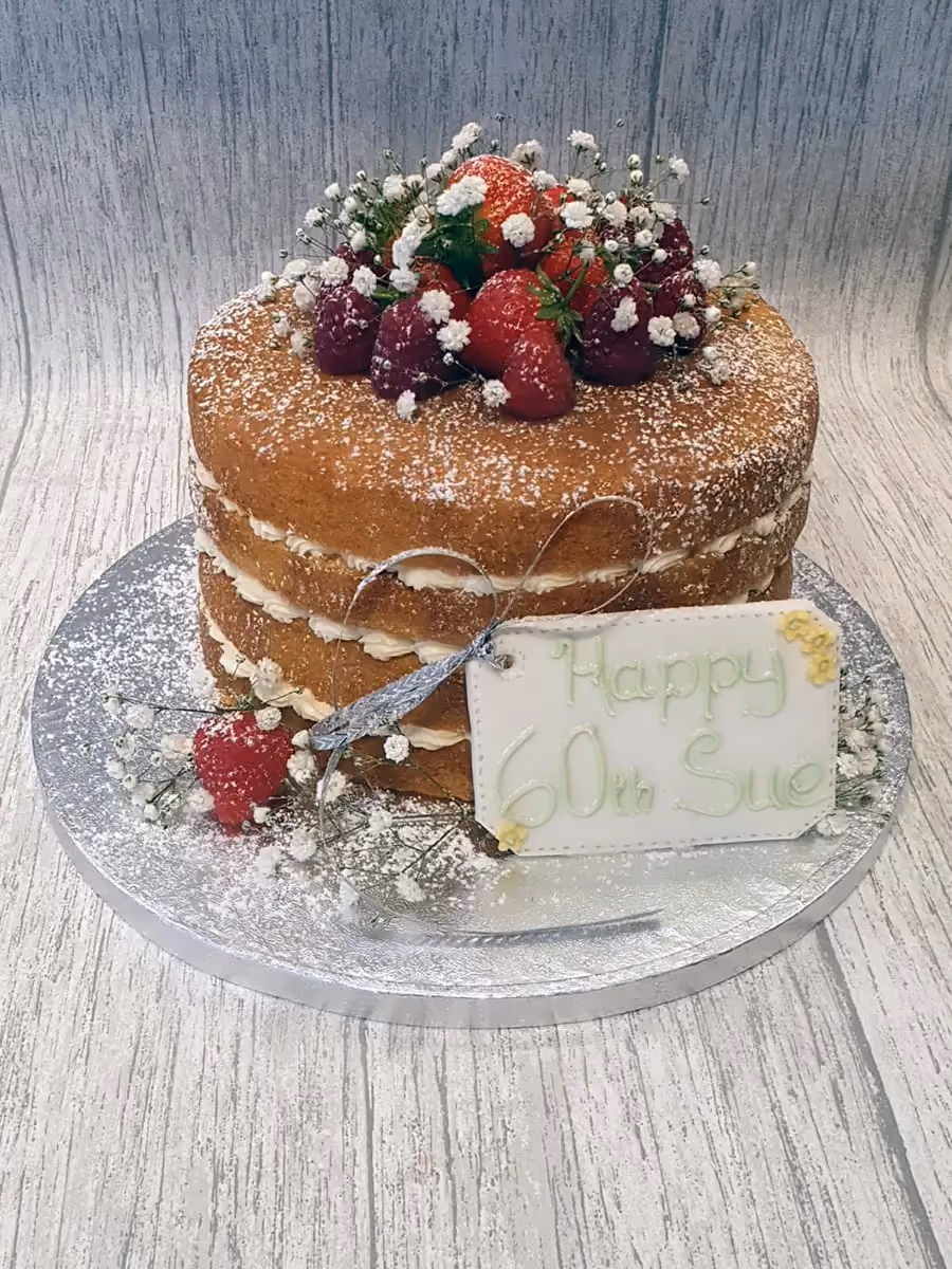 A two-tier naked sponge cake for a 60th birthday, dusted with powdered sugar, and decorated with fresh strawberries, raspberries, white baby's breath flowers, and a 'Happy 60th Sue' message tag.