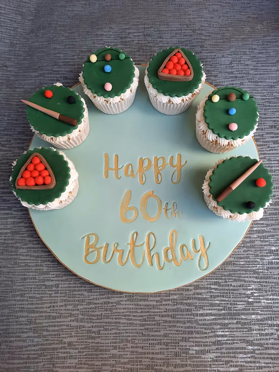 Six snooker or pool-themed cupcakes arranged on a round light green cake board with 'Happy 60th Birthday' written in gold. The cupcakes are decorated with green fondant, miniature cues, colored balls, and racks of red balls.