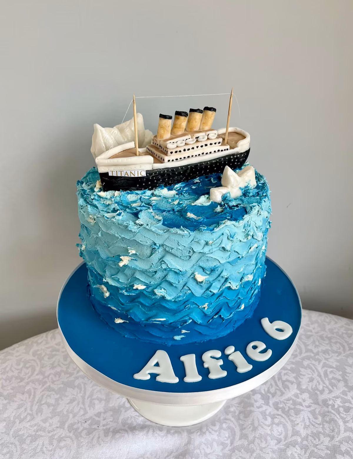 A Titanic themed 6th birthday cake for Alfie with a model ship sailing on textured blue buttercream waves and a dark blue board.
