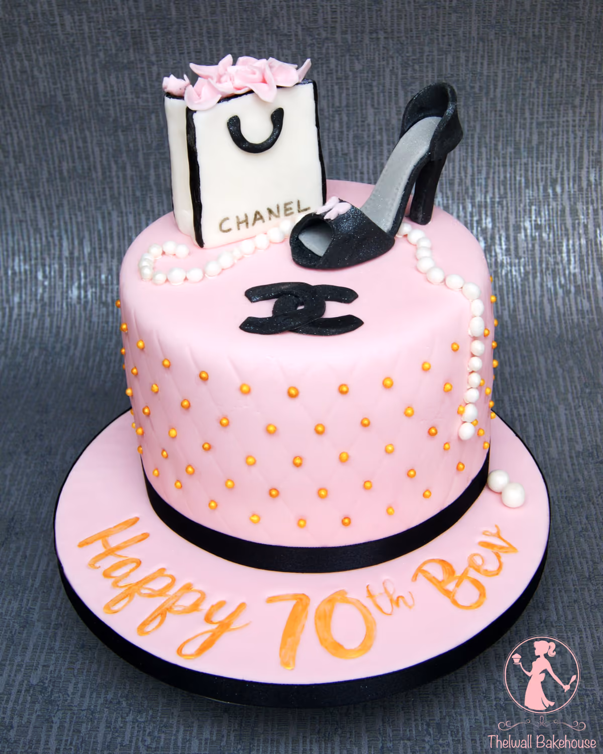 A two-tier pink 70th birthday cake with a Chanel theme, featuring a white shopping bag with pink flowers, a black high-heel shoe, pearl strands, and a quilted pattern with gold beads. The base of the cake reads 'Happy 70th Bev'.