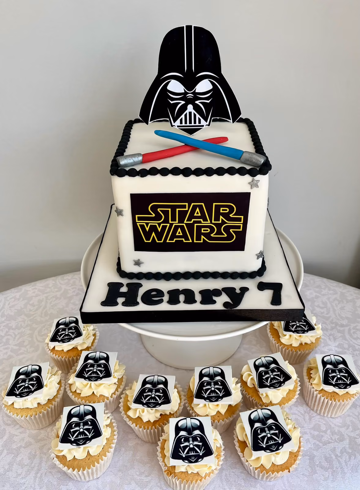 A square Star Wars 7th birthday cake for Henry with a Darth Vader topper and crossed lightsabers, surrounded by matching Darth Vader cupcakes.