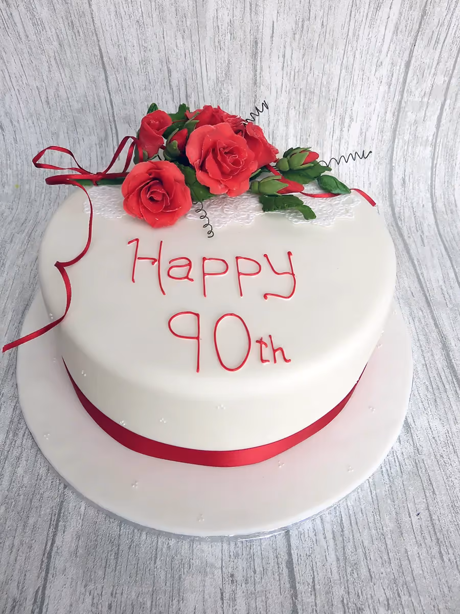 A round white fondant cake for a 90th birthday, decorated with a bouquet of red sugar roses, a red ribbon around the base, and 'Happy 90th' written in red icing.