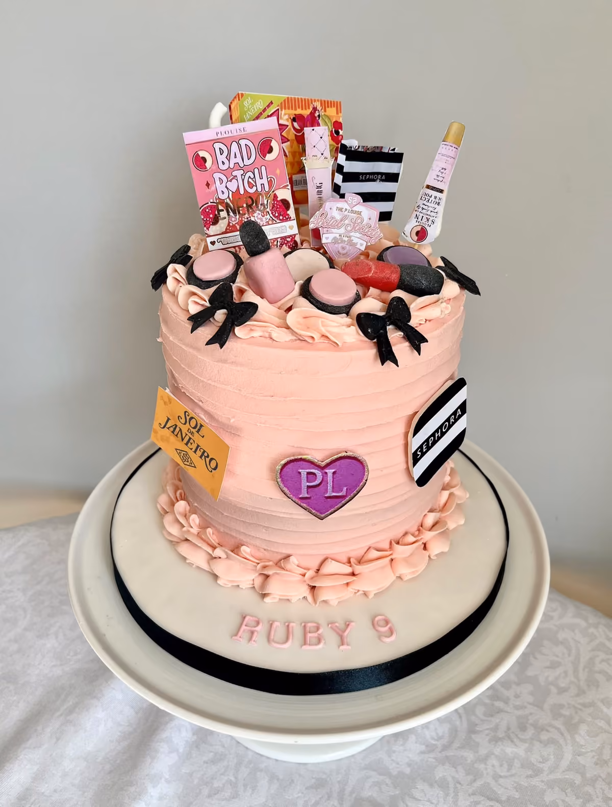 A pink 9th birthday cake for Ruby with black bows, a 'PL' heart and a topper of makeup and skincare products.