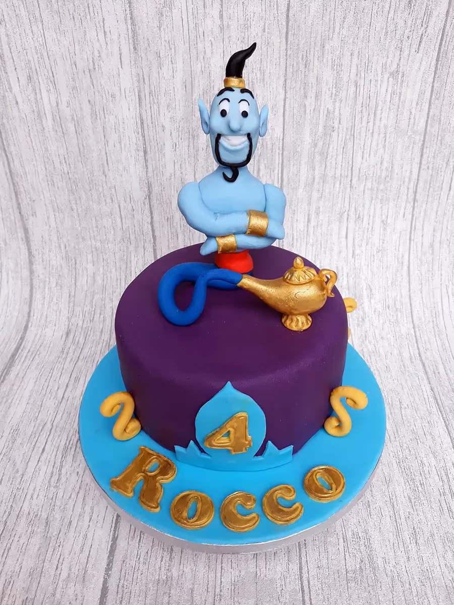 An Aladdin-themed birthday cake featuring a blue Genie figure with crossed arms, a golden magic lamp, a number 4, and the name 'Rocco' on a purple cake with a light blue base.