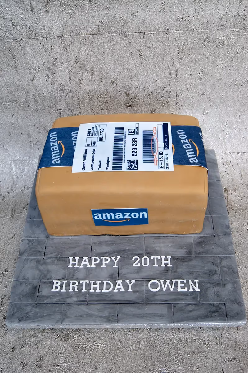 A novelty birthday cake shaped like a brown Amazon package with blue Amazon tape and a shipping label, sitting on a grey tiled base inscribed with 'Happy 20th Birthday Owen'.
