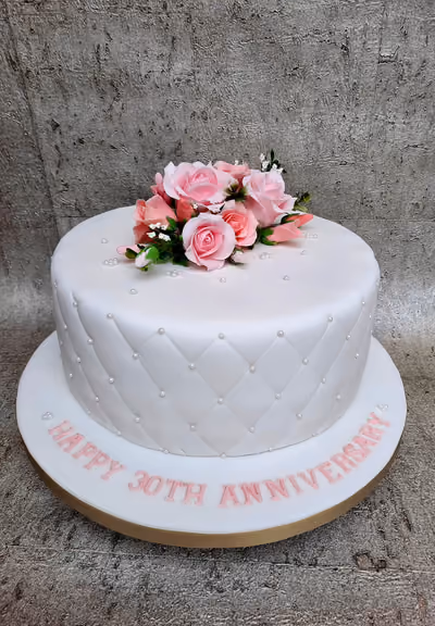 A white, single-tier 30th anniversary cake with a quilted pattern and pearl details, topped with a bouquet of pink sugar roses and inscribed with 'Happy 30th Anniversary' in pink letters on the cake board.